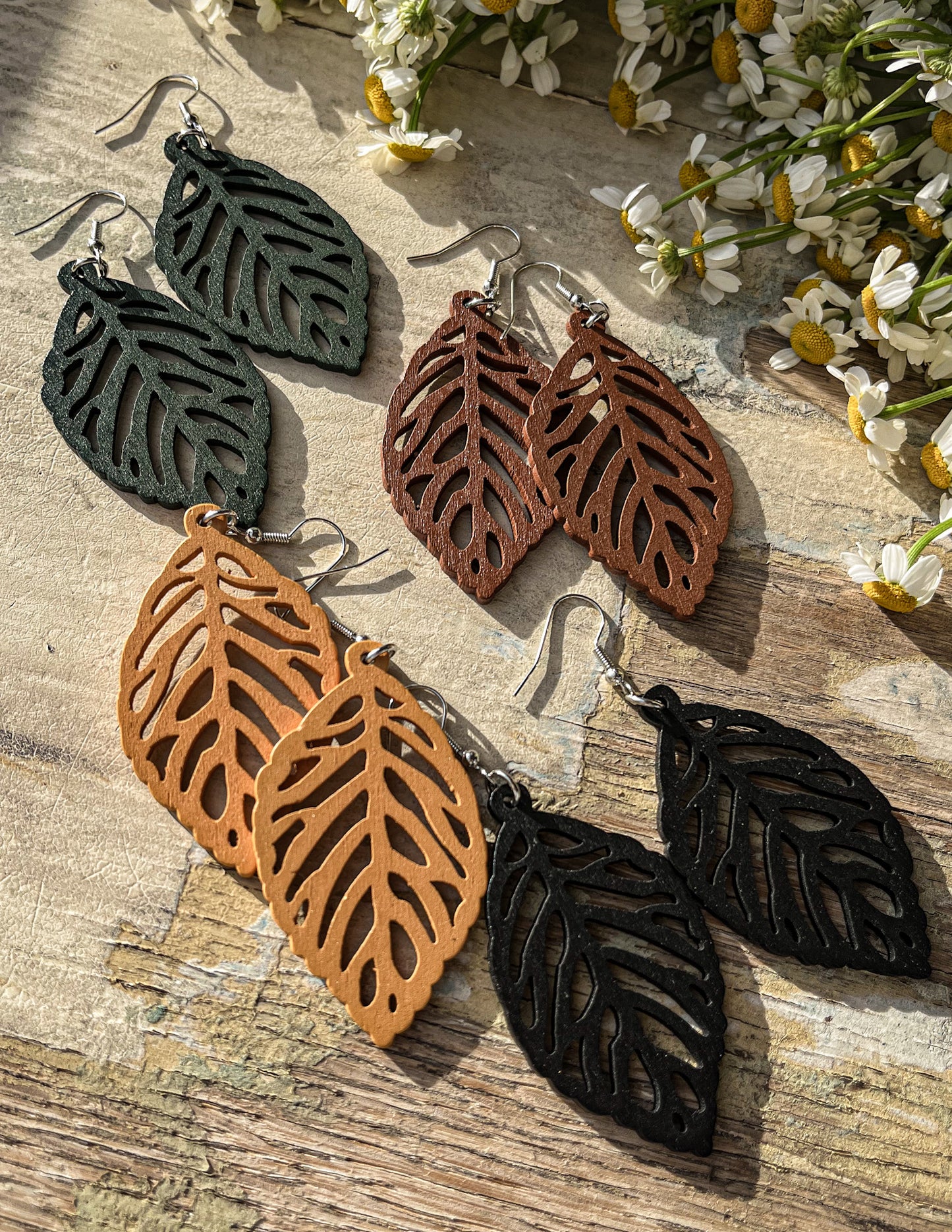 Laser-cut leaf earrings in dark green brown tan and black featuring intricate cut-out patterns artfully arranged on a weathered wooden surface beside scattered white daisies in bright sunlight