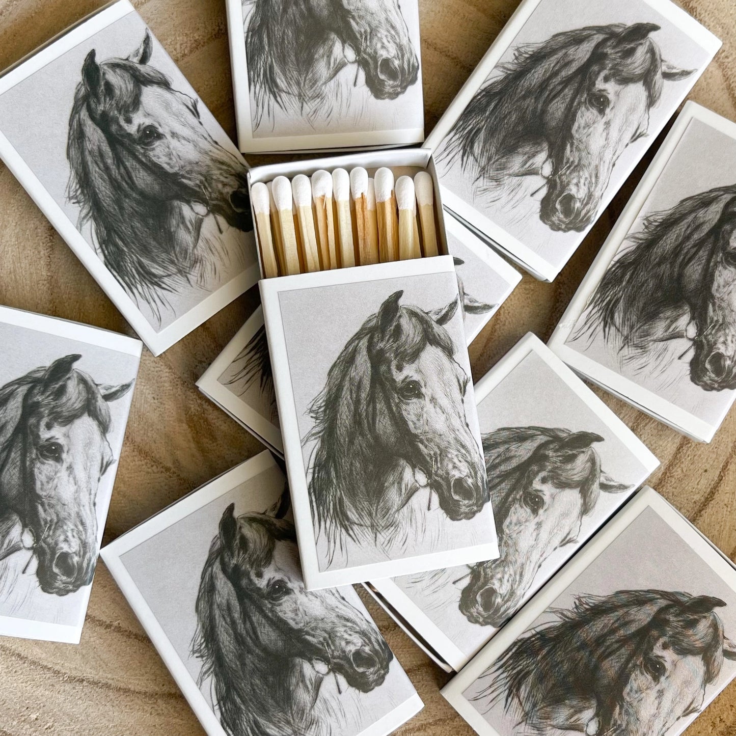 Multiple matchboxes featuring black and white charcoal-style horse head drawings are scattered on a light wooden surface with one open box revealing white-tipped matches