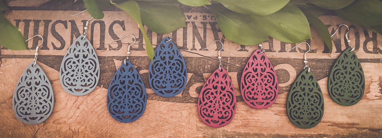 Photograph of six wooden teardrop earrings in light blue, deep blue, hot pink, and olive green, each featuring intricate laser-cut filigree patterns, suspended from silver hooks against a rustic background with faded "SUNSHINE" text and fresh green leaves