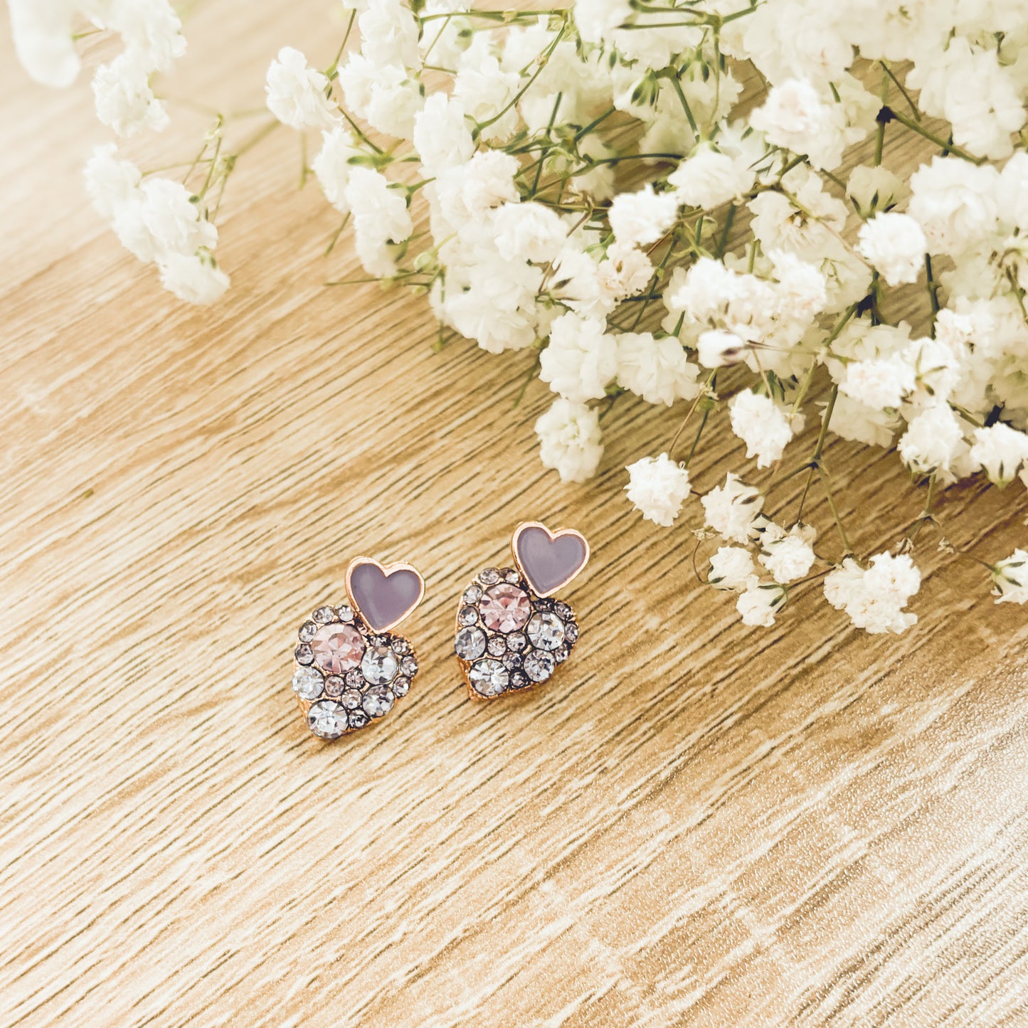 Pair of heart-shaped rose gold earrings featuring sparkling clear and light pink gemstones with small lavender enamel hearts on a light wood surface alongside delicate white baby's breath flowers