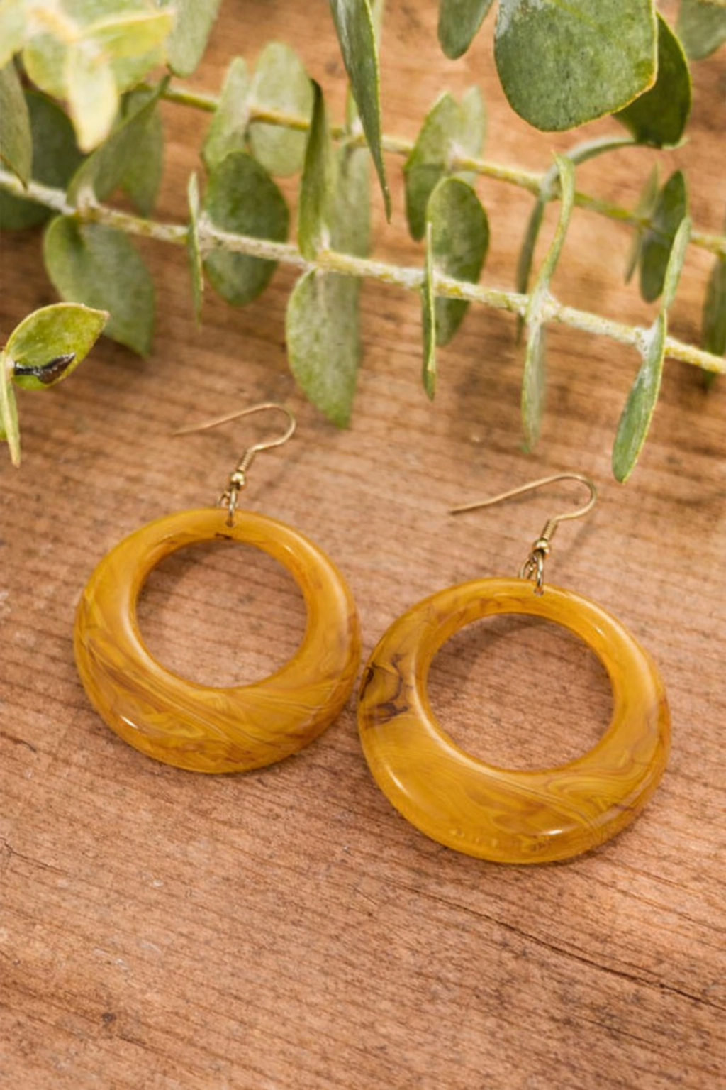 A pair of marbled mustard yellow resin hoop earrings with gold-toned fishhook findings rests on a rustic wooden surface with soft green eucalyptus leaves in the background