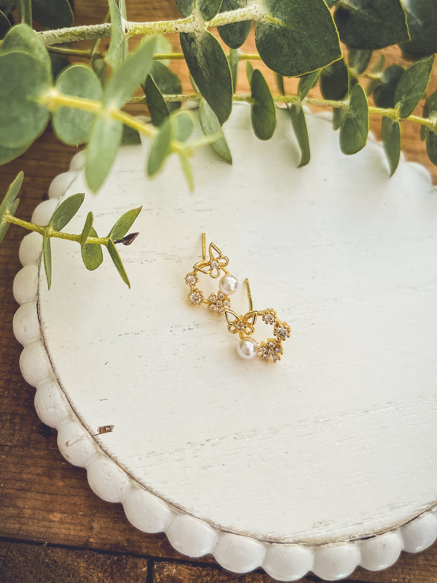 Photograph of a pair of gold-toned butterfly and floral cluster earrings on a white scalloped tray each adorned with clear gemstones and a single pearl accented by soft green eucalyptus leaves and a rustic wooden surface