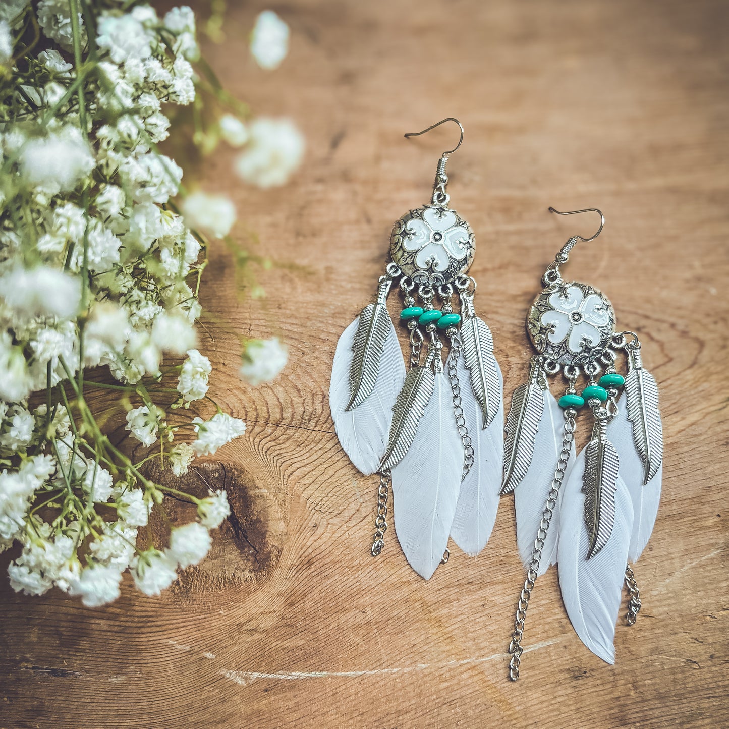 Product photography featuring a pair of bohemian silver earrings with white feathers, turquoise beads, and a central white floral design, resting on a rustic wooden surface beside soft white baby's breath flowers