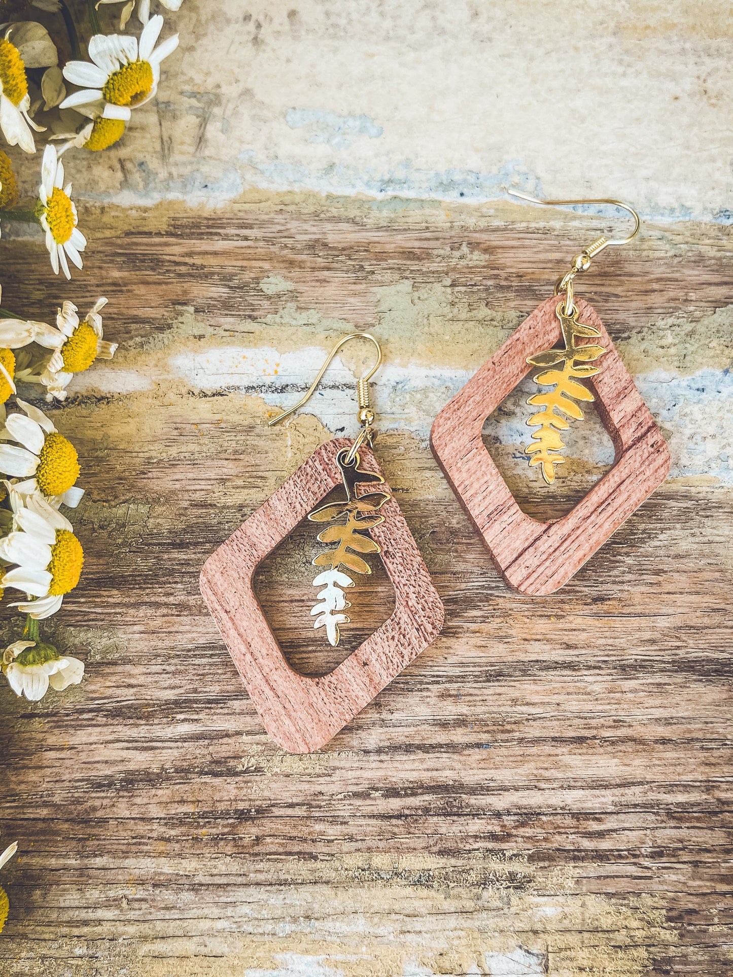 Handcrafted diamond-shaped light brown wood and golden fern leaf earrings displayed on a weathered wooden surface with dried white and yellow daisies