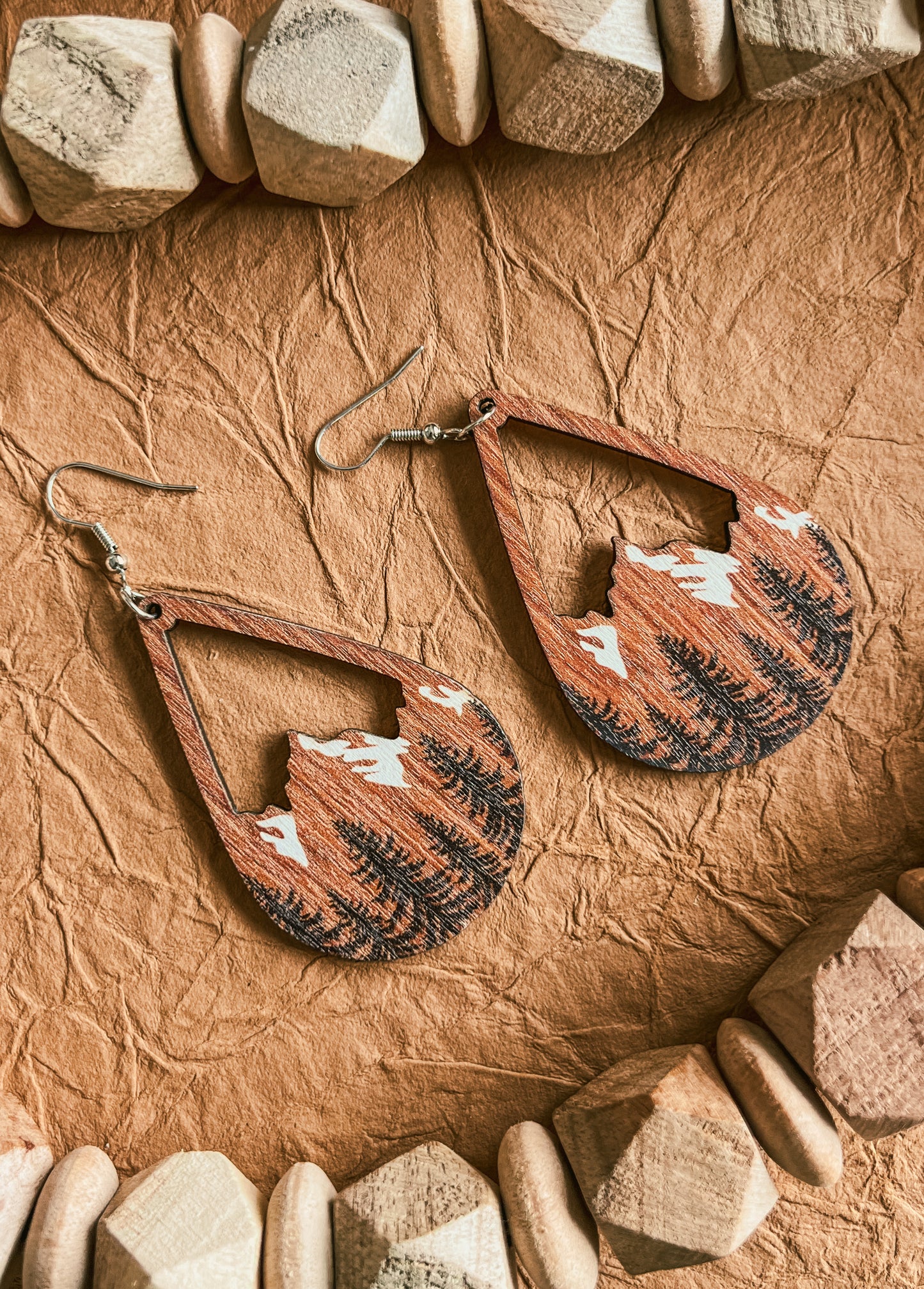 Pair of handcrafted wood teardrop earrings depicting etched mountain and pine forest designs with white snow caps, presented on crumpled tan paper bordered by light wooden beads