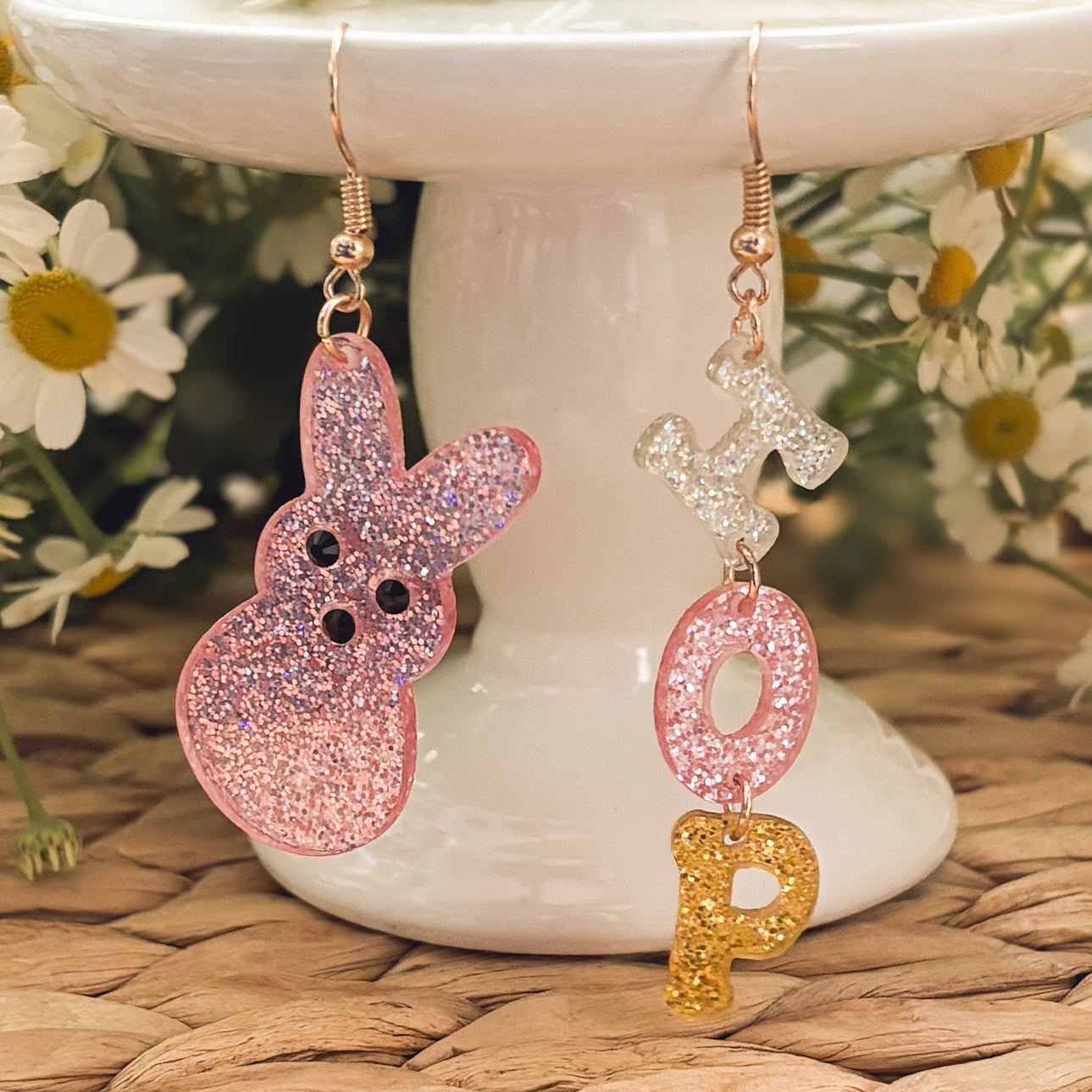 Acrylic resin earrings featuring rose gold hooks, one a pink glitter Peeps bunny with black eyes and the other spelling "HOP" with white, pink, and gold glitter letters, displayed on a white cake stand against daisies and a woven mat