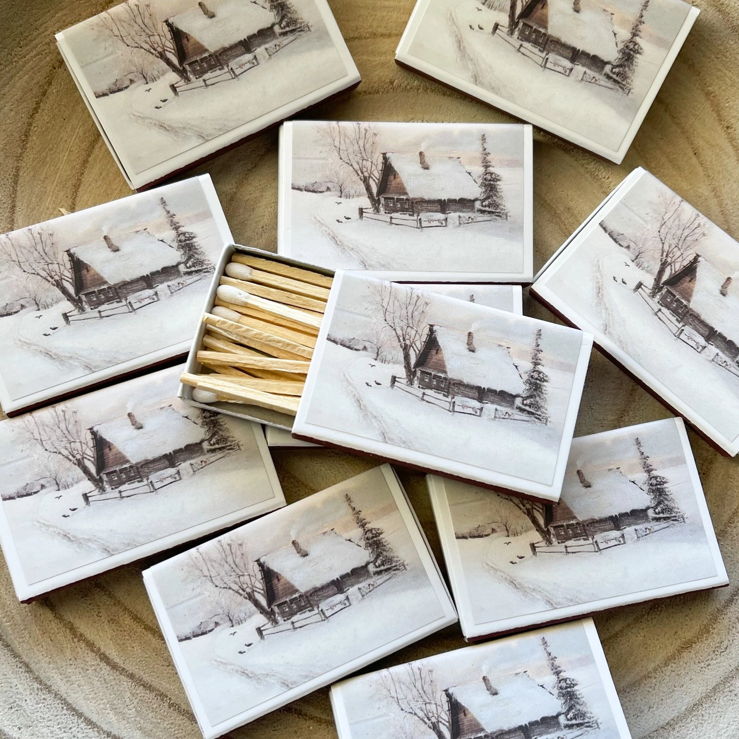Photograph of multiple matchboxes arranged on a light wooden surface, each featuring a winter scene of a snow-covered log cabin with a smoking chimney, bare trees, a tall evergreen, a snowy path, and small birds, with one box open revealing numerous light wooden matches