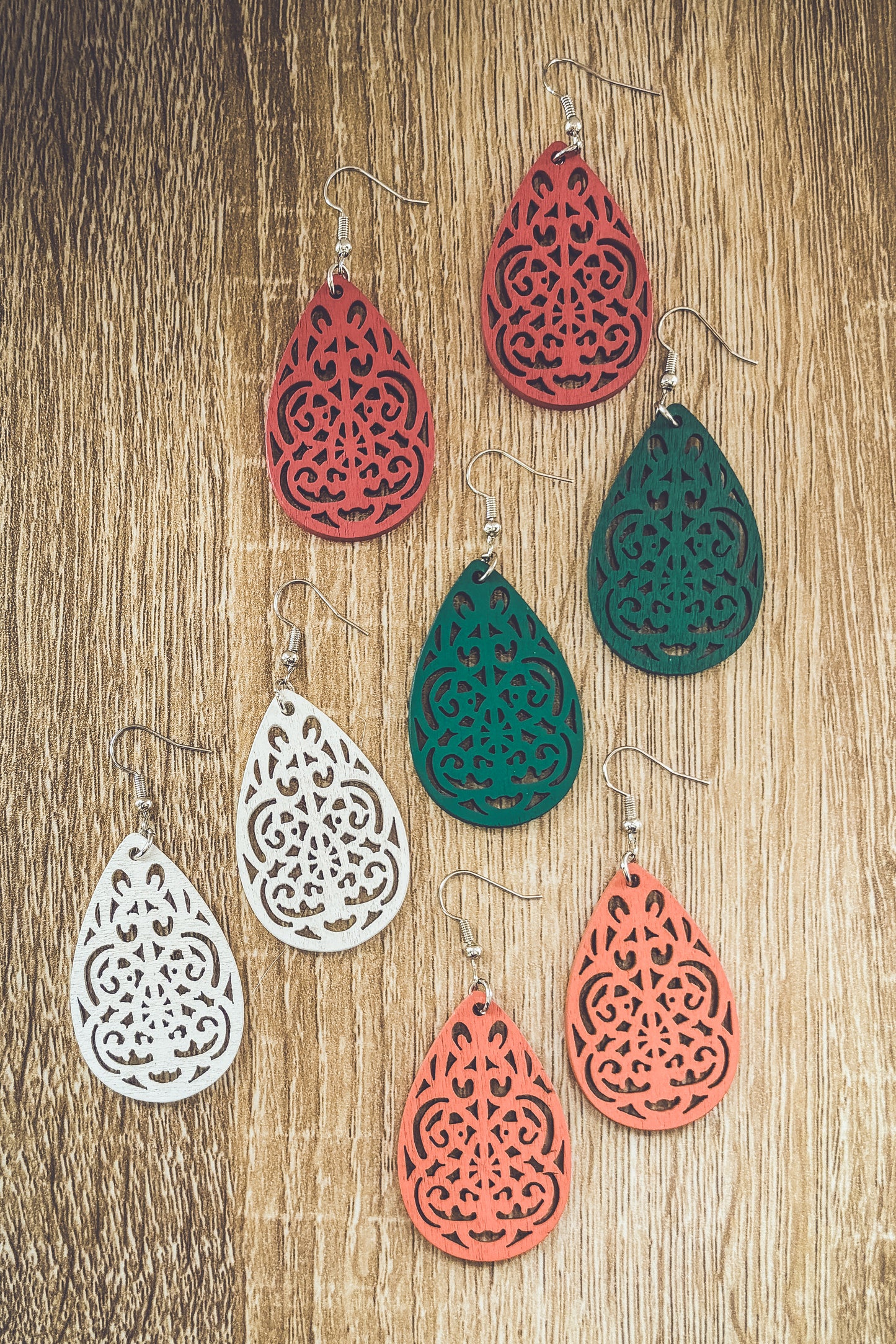 Handcrafted wooden teardrop earrings featuring intricate laser-cut patterns in red, green, white, and coral displayed on a natural wood grain surface