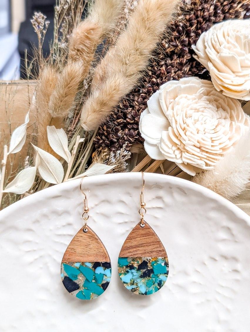 Photograph of a pair of artisan teardrop earrings featuring polished wood tops and resin bottoms with turquoise, light blue, black flakes and gold glitter, resting on a white textured dish against a background of dried pampas grass and sola wood flowers