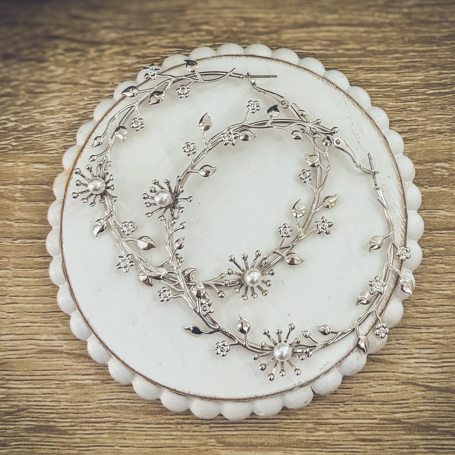 A pair of silver-toned hoop earrings with a delicate floral vine design adorned with faux pearls and tiny clear rhinestones, resting on a white scalloped coaster against a rustic wooden background