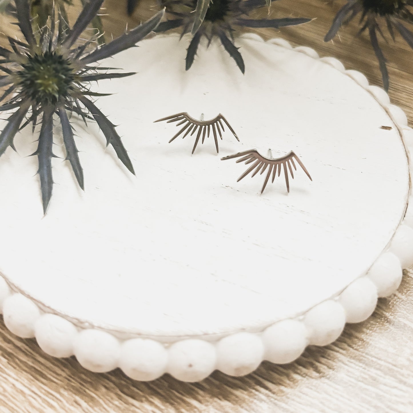 Silver-toned sunburst stud earrings on a white beaded wooden tray, beside a spiky blue eryngium thistle