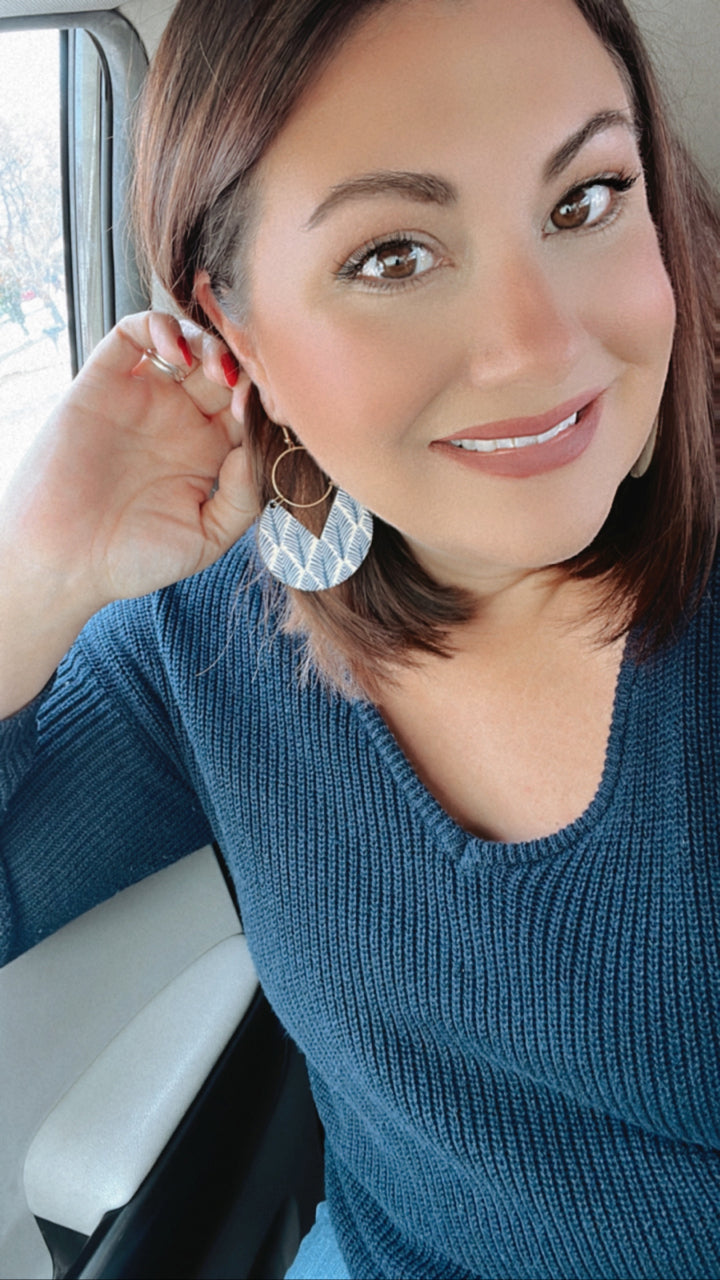 A smiling woman with brown hair and warm makeup in a dark blue ribbed knit sweater features a blue and white patterned semicircular earring and red nail polish