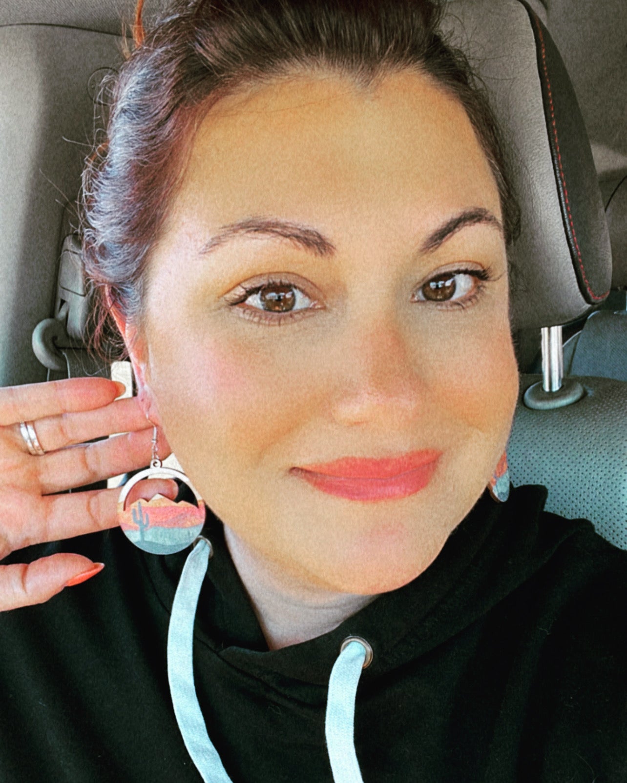 Photograph of a smiling woman in a car wearing coral lipstick and a black hoodie, showcasing a unique dangle earring featuring pastel desert mountains and a saguaro cactus design