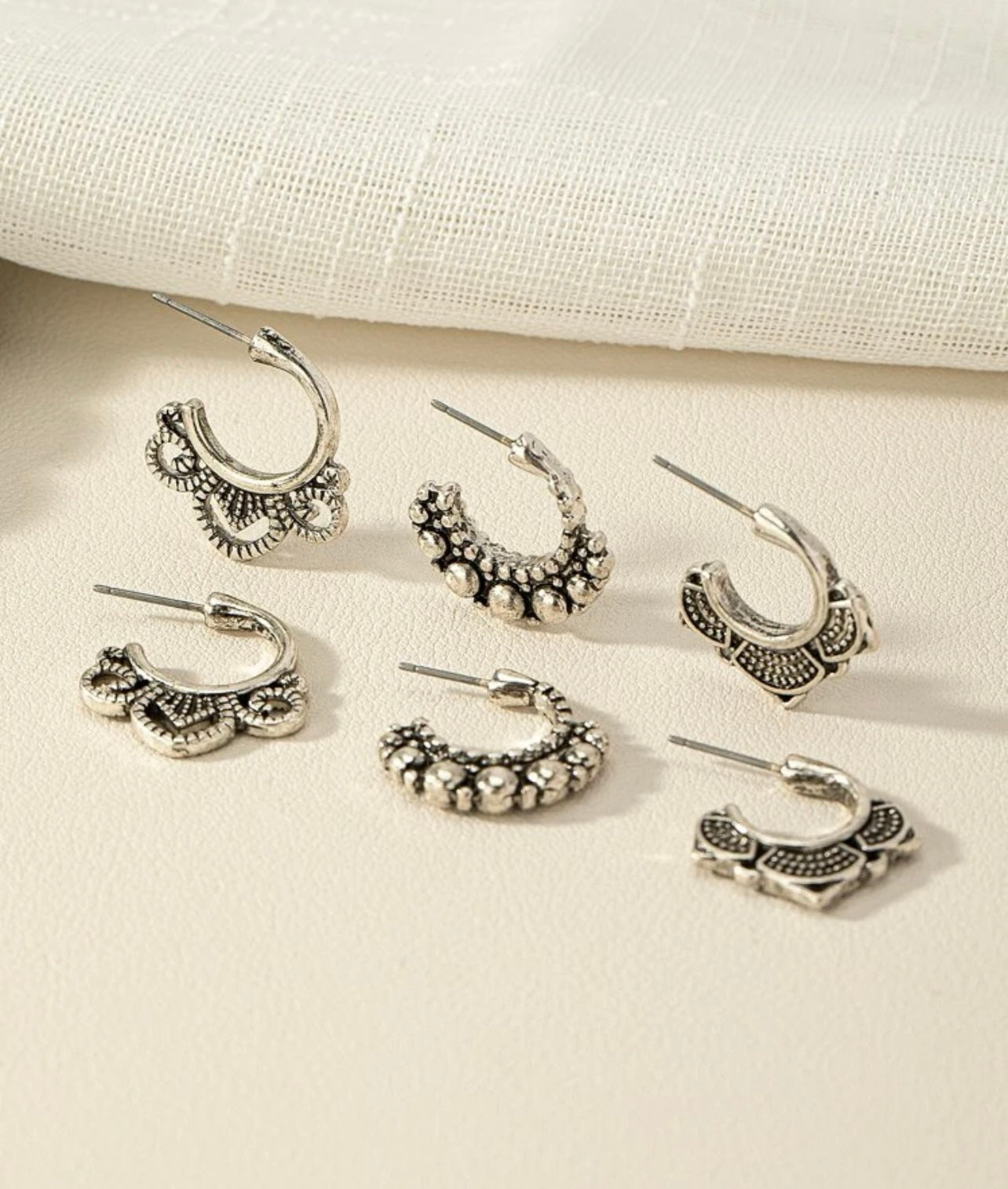 A set of six antique silver stud hoop earrings displayed on a light cream surface featuring three distinct bohemian-inspired designs with intricate filigree, textured beaded borders, and stylized floral patterns
