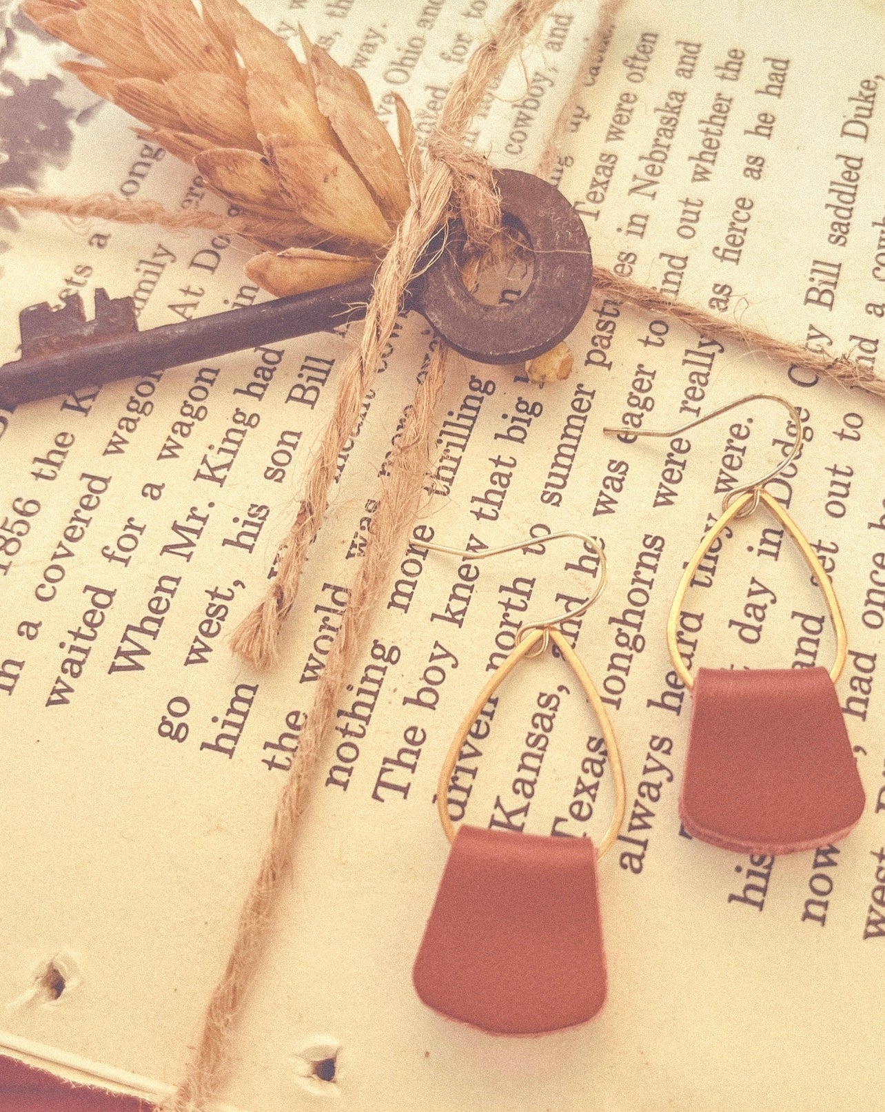A rustic flat lay featuring brown leather drop earrings, an antique skeleton key, and dried wheat tied with jute twine, all arranged on an old book page with Western-themed text mentioning Kansas and Texas