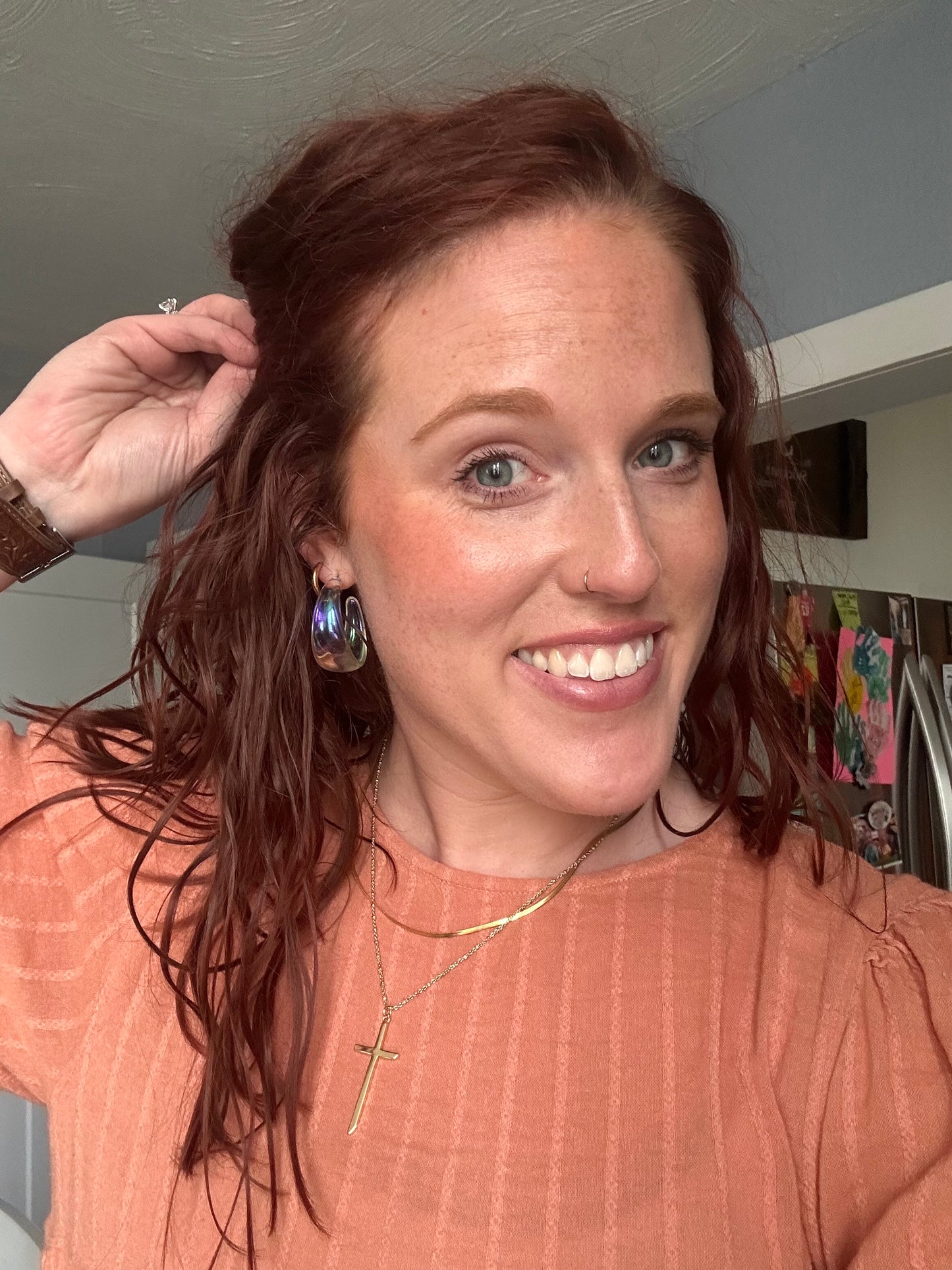 Photograph of a smiling red-haired woman with freckles, her hand in her hair, wearing iridescent earrings and a gold cross necklace, in a home kitchen