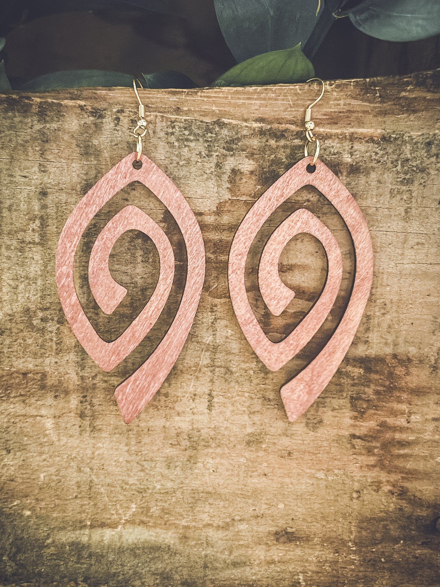 A pair of soft pink wooden earrings featuring an abstract leaf shape with spiral cutouts hangs from gold-toned hooks against a textured rustic wood plank, with dark green foliage subtly visible above