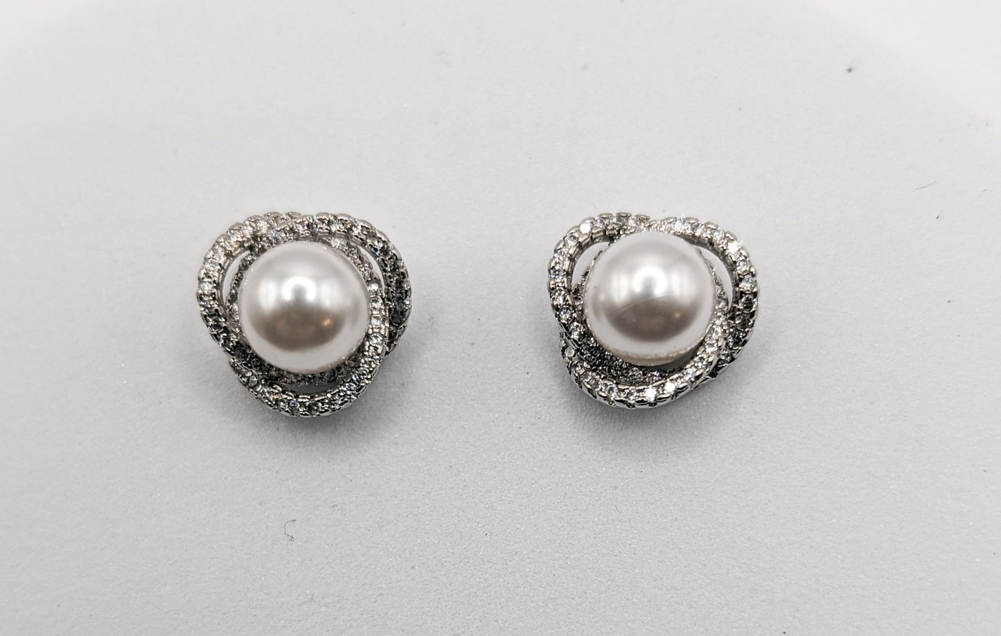 A pair of elegant stud earrings featuring lustrous white pearls encircled by a sparkling twisted pavé setting of brilliant clear cubic zirconia stones