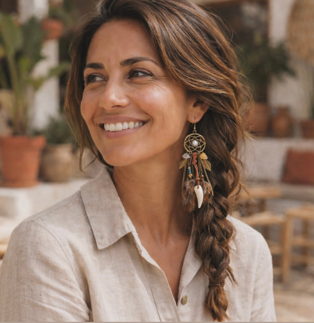 A smiling woman with long braided brown hair wears a distinctive bohemian dreamcatcher earring with feathers and a white tusk charm in a warm outdoor setting with blurred terracotta pots