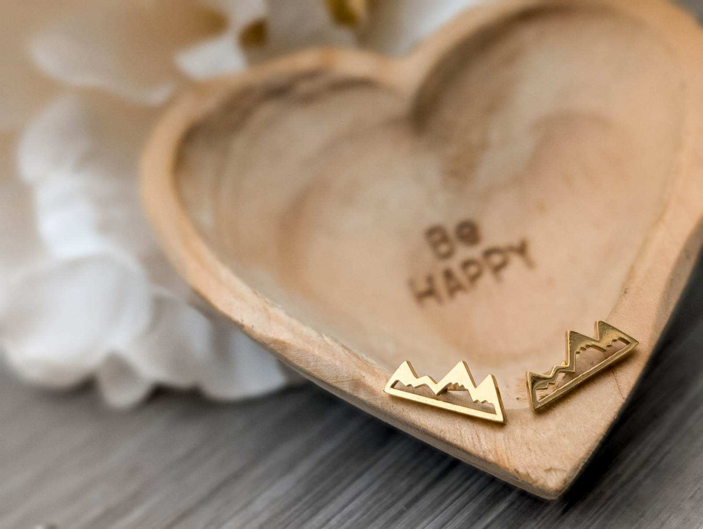 Photograph of two gold mountain range stud earrings on a heart-shaped wooden dish etched with 'Be Happy' against a blurred background of white flower petals and gray wood