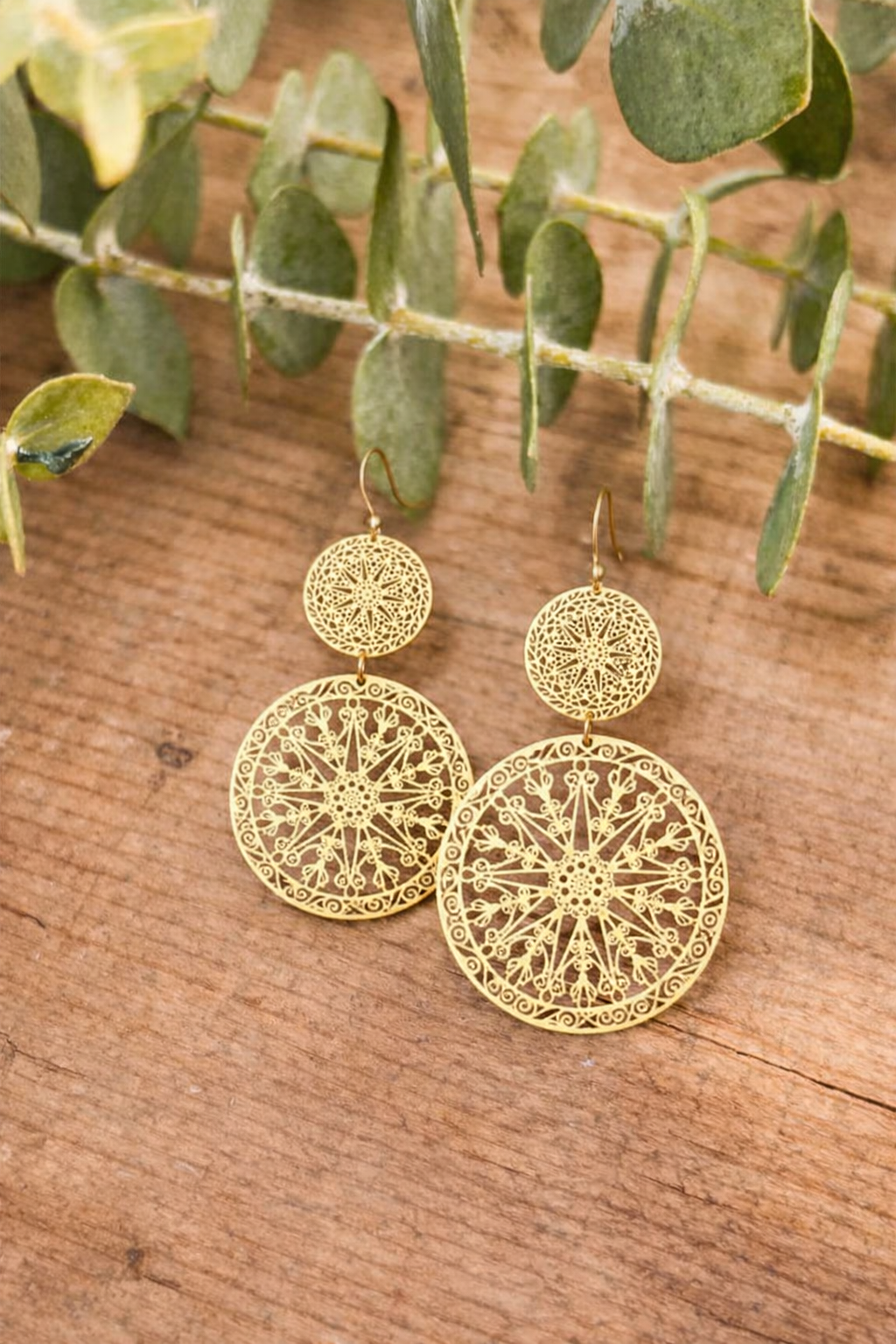 Gold earrings two ornate gold-tone dangle earrings with intricate filigree star and floral patterns displayed on a rustic wooden background with blurred green eucalyptus foliage