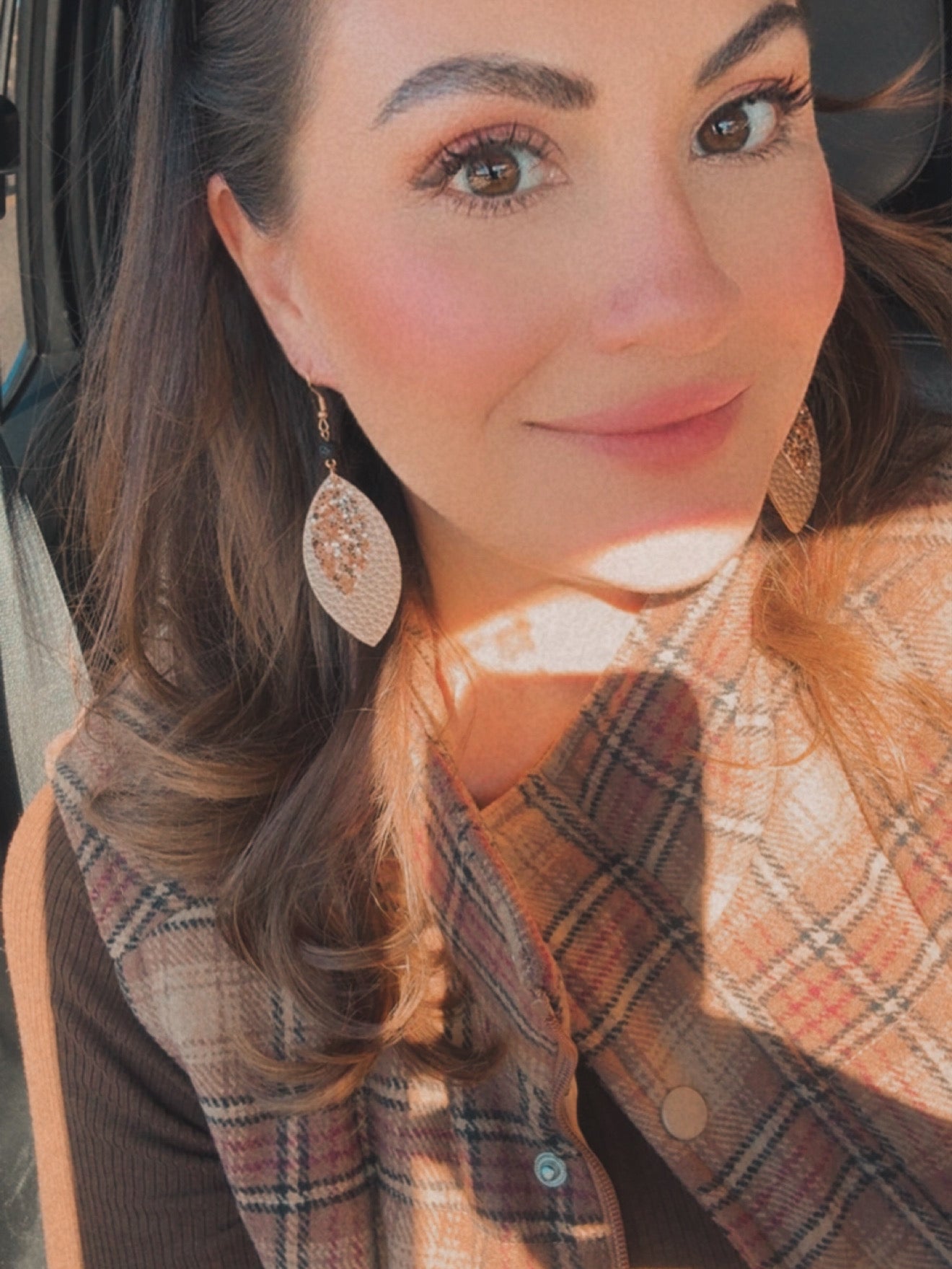 Photograph of a smiling woman with brown eyes and soft sunlight dappling her brown wavy hair, wearing a brown and beige plaid vest and prominent glittery leaf-shaped earrings