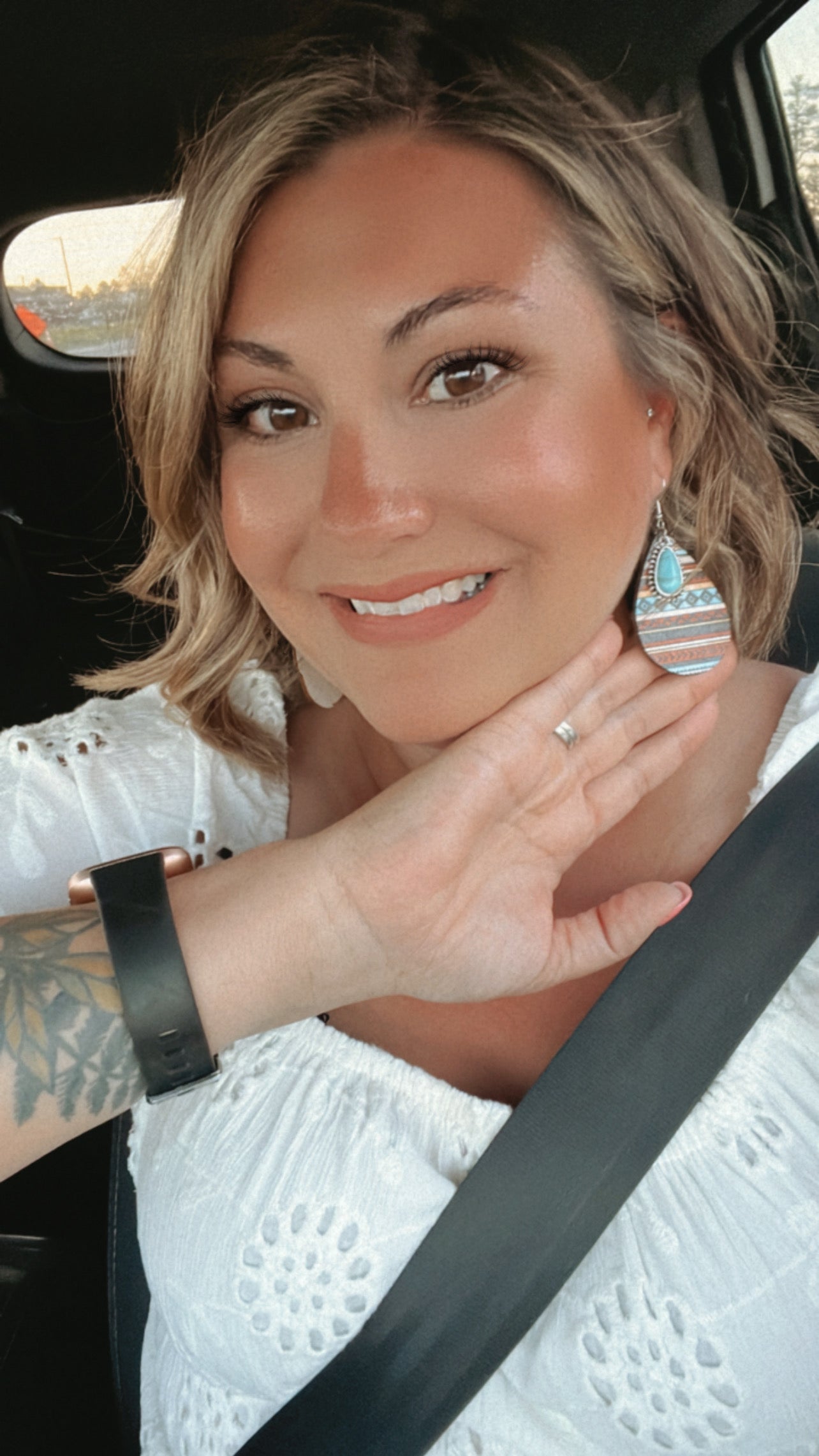 Photograph of a smiling blonde woman in a car wearing a white eyelet top, a distinctive tribal turquoise earring, and a visible arm tattoo