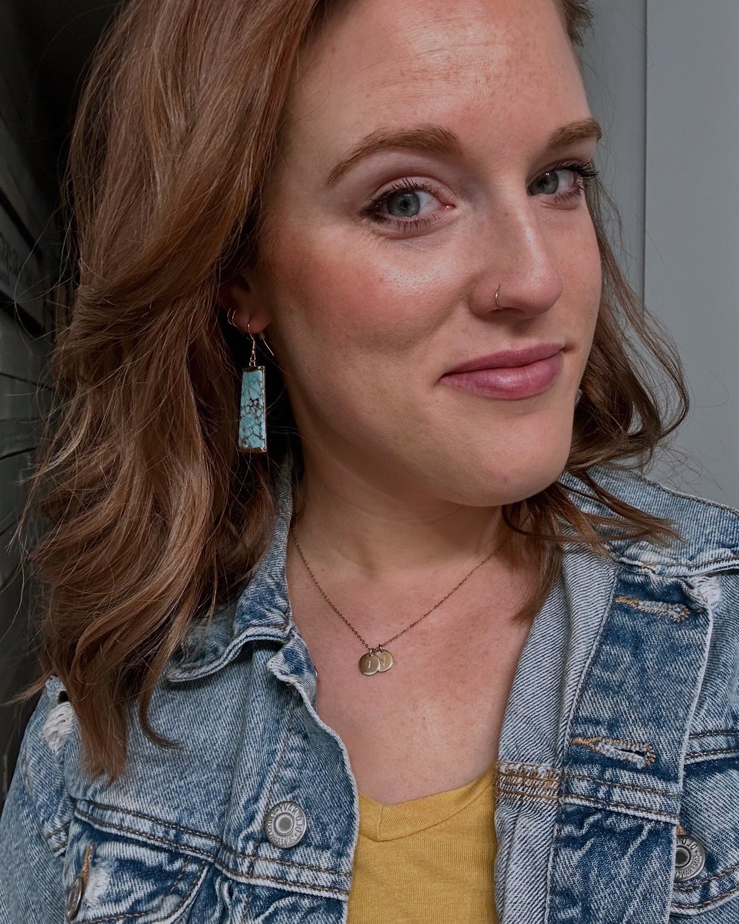 Close-up portrait of a woman with wavy auburn hair and a slight smile, wearing rectangular turquoise and black matrix earrings, a subtle gold nose ring, and a distressed denim jacket over a mustard yellow top