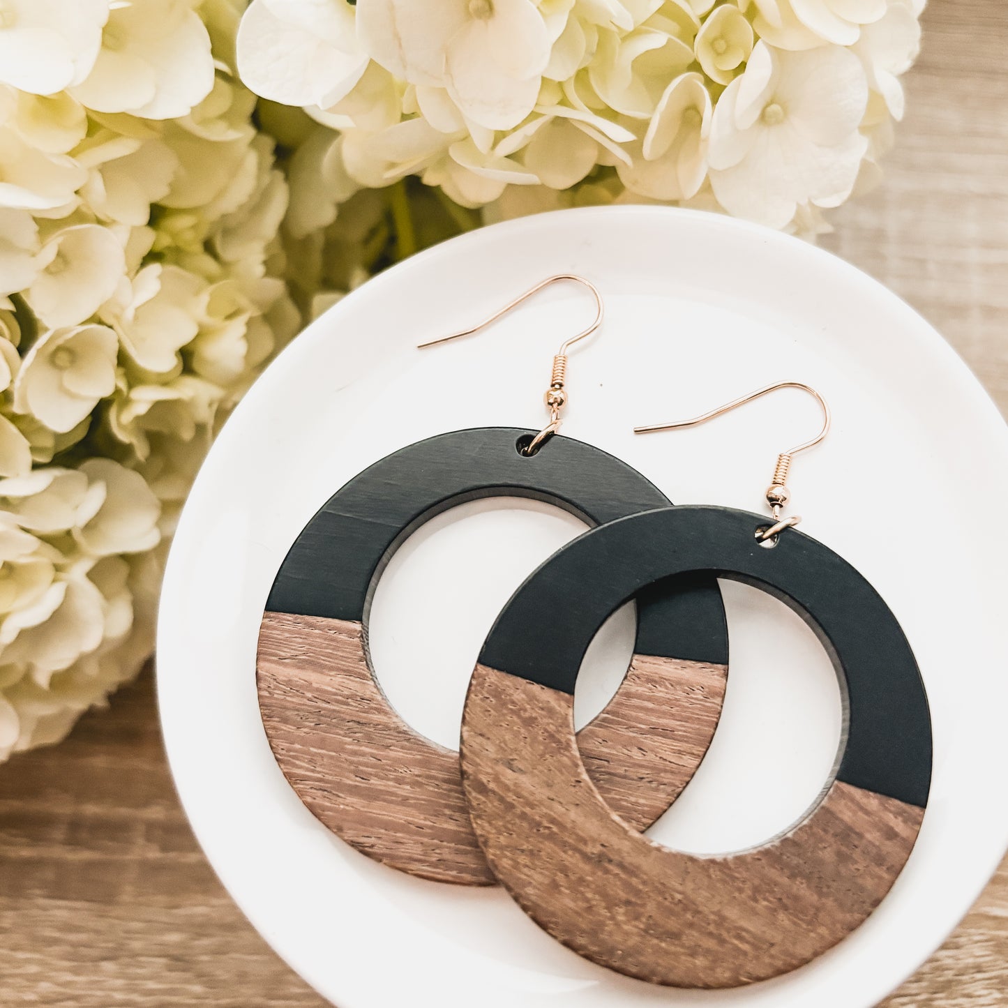 A pair of circular two-tone earrings featuring matte black and natural wood sections with rose gold hooks resting on a white ceramic dish, blurred white hydrangea flowers and a light wooden surface in the background