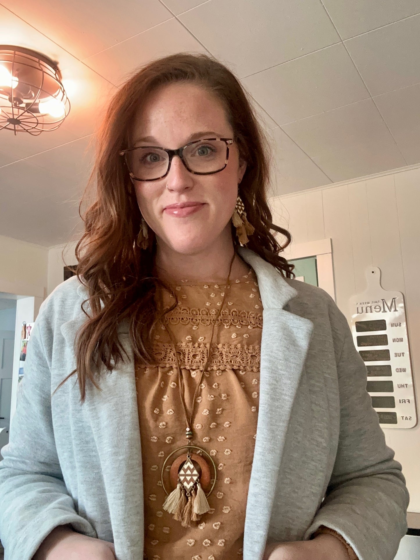 A smiling woman with auburn hair and glasses wears a light grey blazer over a mustard yellow swiss dot blouse with lace trim, featuring a large boho tassel necklace and matching earrings, with an industrial caged ceiling light and a weekly menu board visible in the background