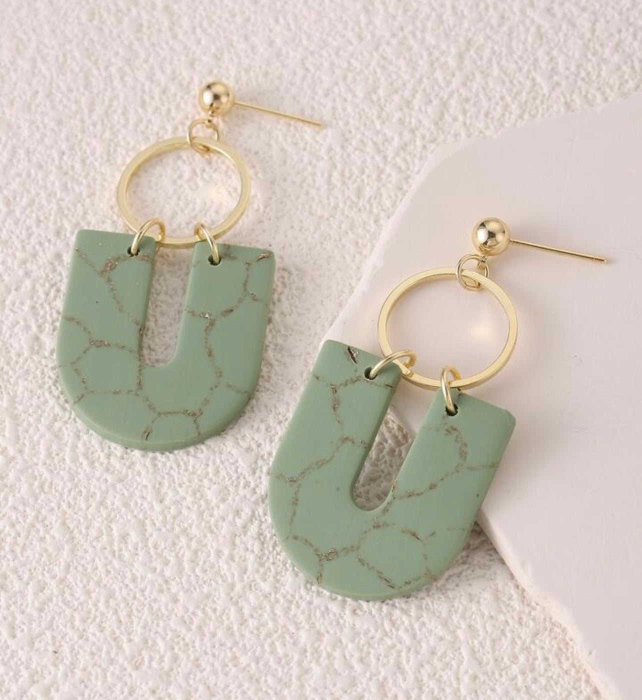 Pair of modern dangle earrings featuring U-shaped faux malachite green pendants with gold veining, suspended from gold-tone hoops and ball studs, presented against a subtly textured white surface
