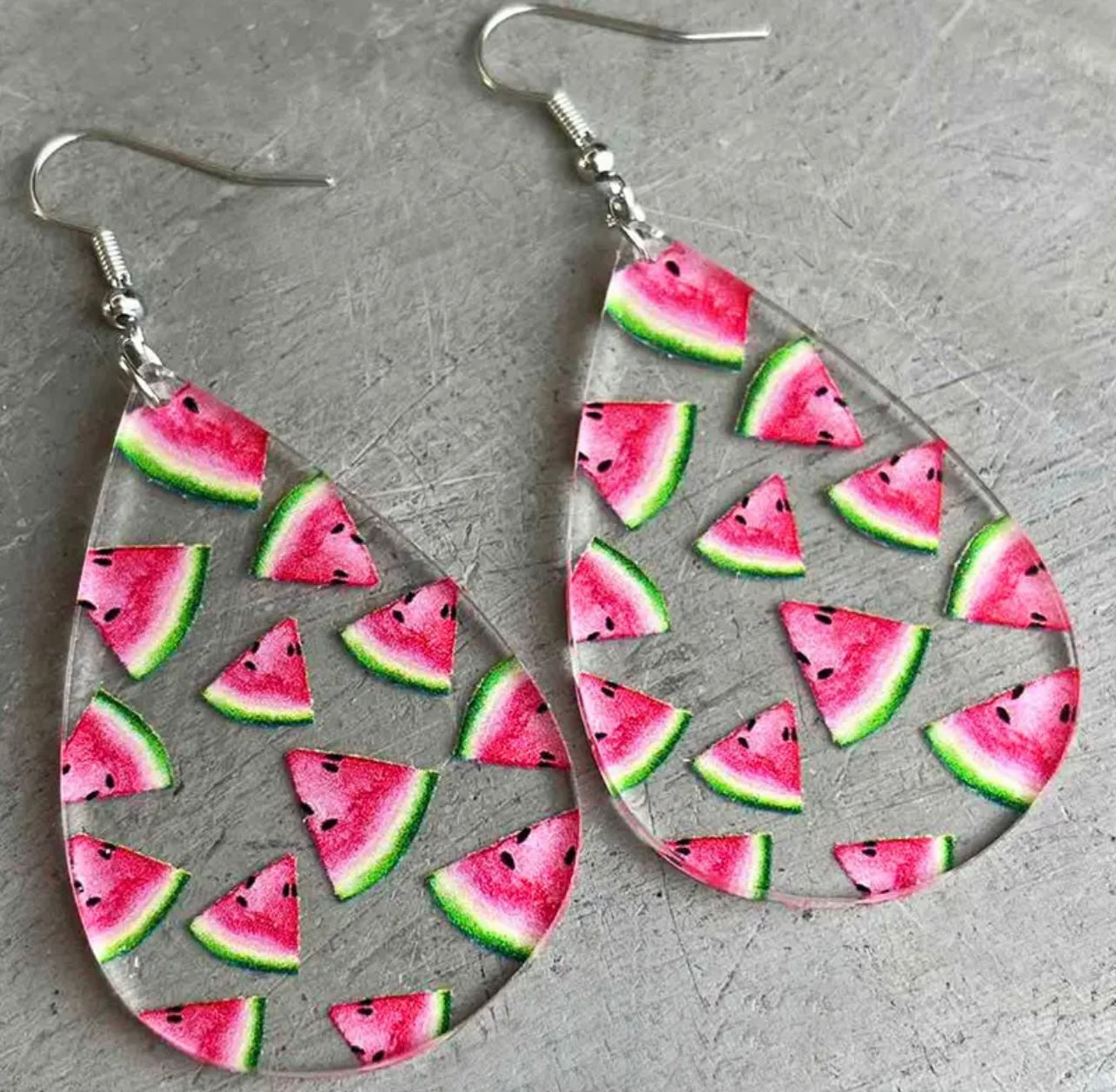 Clear acrylic teardrop earrings featuring vibrant pink watermelon slices with green rinds and black seeds, suspended by silver hooks on a grey backdrop
