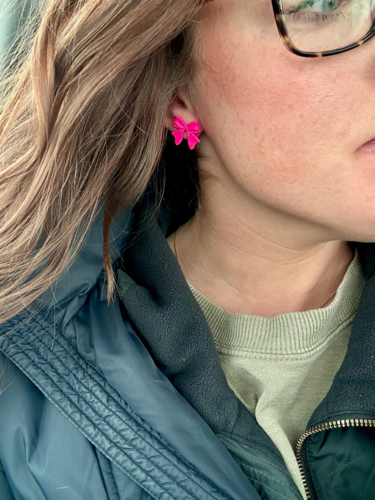 Photograph of a person's side profile with light blonde hair, wearing a vibrant fuchsia pink bow earring, tortoiseshell glasses, and a dark blue jacket