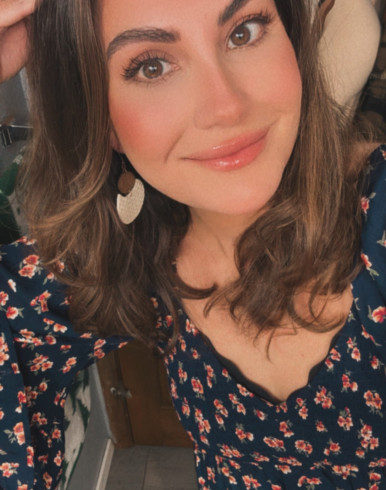Photograph of a smiling woman with glossy lips and dark brown hair wearing a dark blue dress with small red and orange flowers and a textured gold and cream earring indoors
