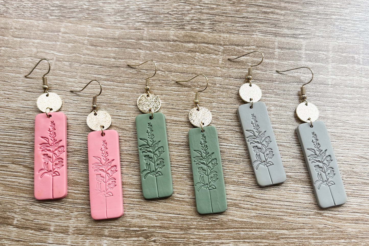 Handmade polymer clay dangle earrings arranged on a wooden surface featuring six rectangular pendants in pink, sage green, and light gray embossed with delicate botanical designs and connected to hammered gold-tone circular studs