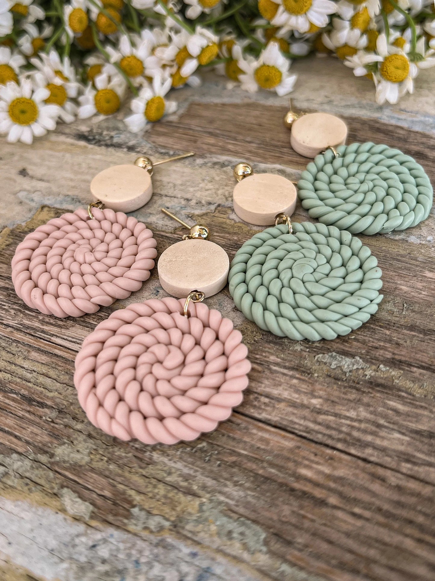 Handmade polymer clay earrings in pink and mint green with a distinctive spiral woven texture and beige circular studs displayed on a rustic wooden surface with blurred white daisies in the background