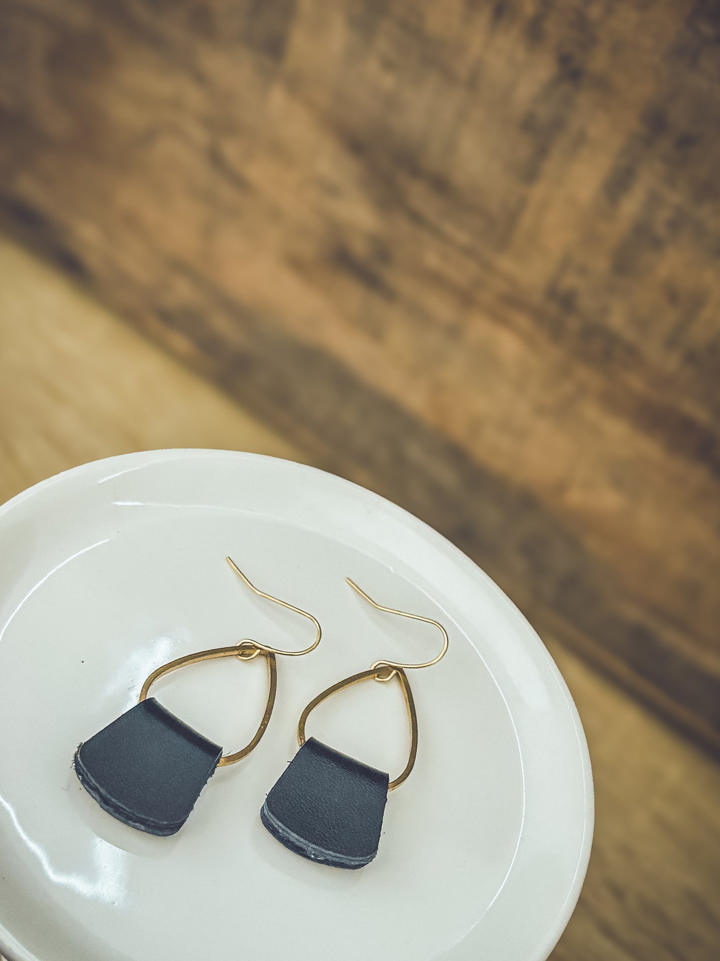 Photograph of two handcrafted earrings featuring dark grey leather pendants suspended from brass teardrop hooks, displayed on a glossy white ceramic plate over a textured wooden surface