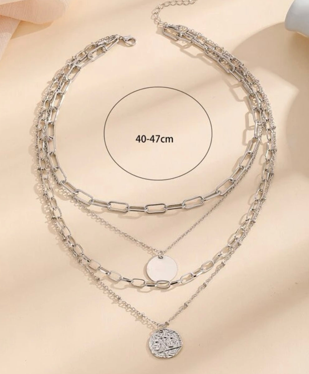 Silver three-layer necklace displayed on a beige background featuring a top chain with thick rectangular links, a middle chain with a plain round disc pendant, and a bottom chain with a textured coin-like pendant, with an overlaid measurement of 40-47cm