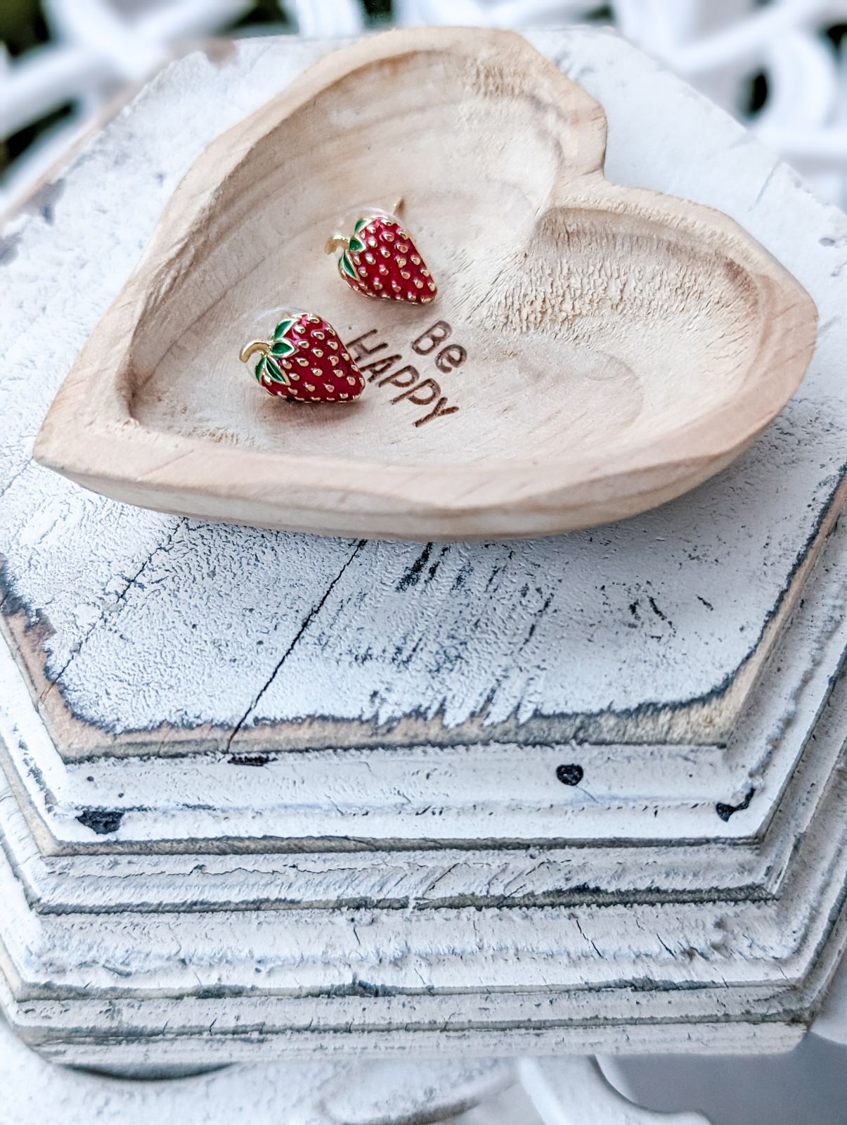 Two vibrant red enamel strawberry stud earrings rest in a light wood heart-shaped dish engraved with "Be HAPPY" on stacked distressed white hexagonal wooden pieces