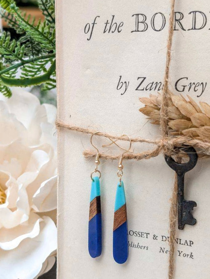 A still life featuring a pair of vibrant blue, turquoise, and wood teardrop earrings hanging from rustic twine over a vintage book titled of the BORDER by Zane Grey, accented by a black skeleton key, dried wheat, and soft white florals