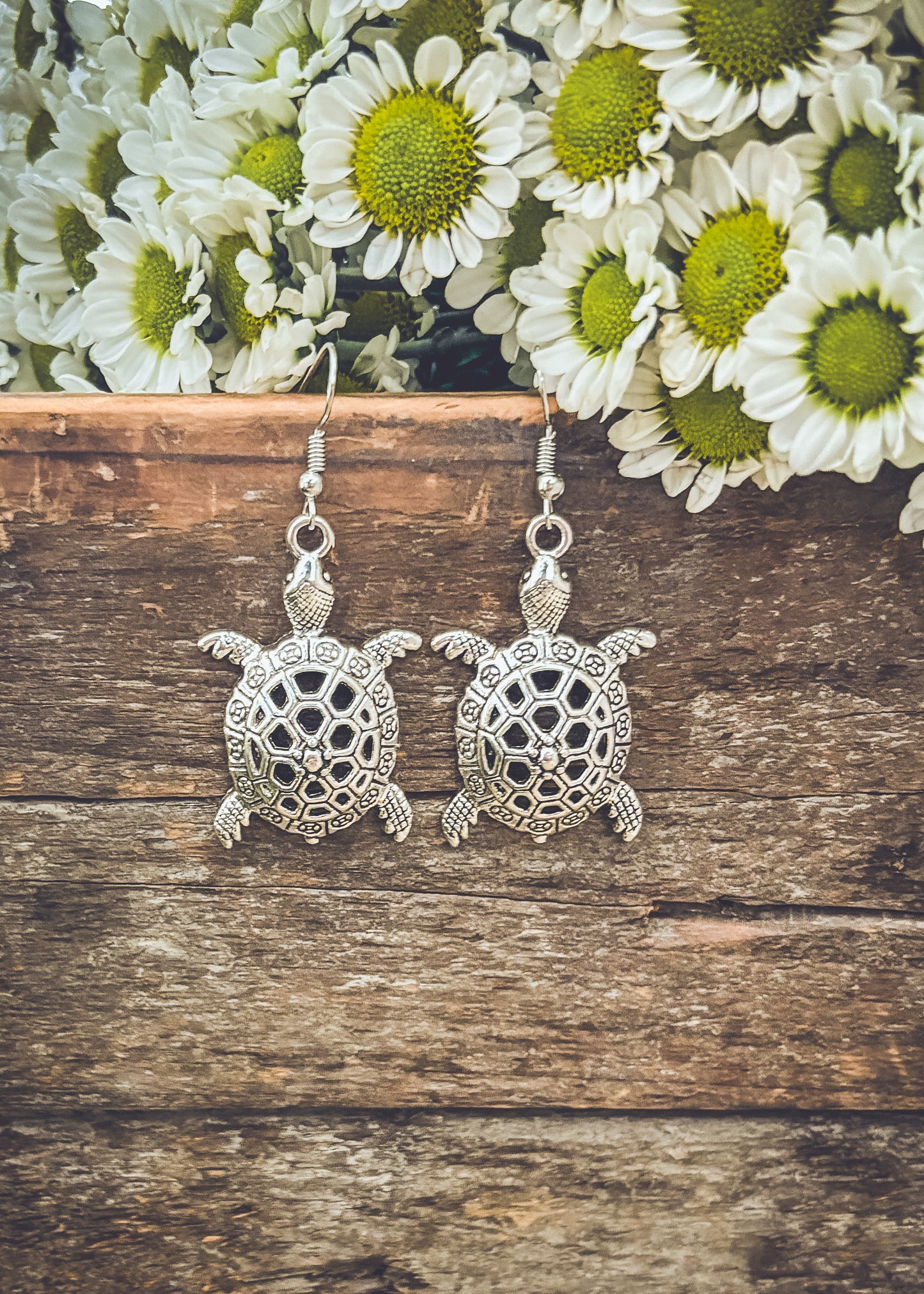 Photograph of a pair of silver turtle earrings with intricate shell designs resting on a rustic wooden surface with a cluster of white and green chrysanthemums blooming above