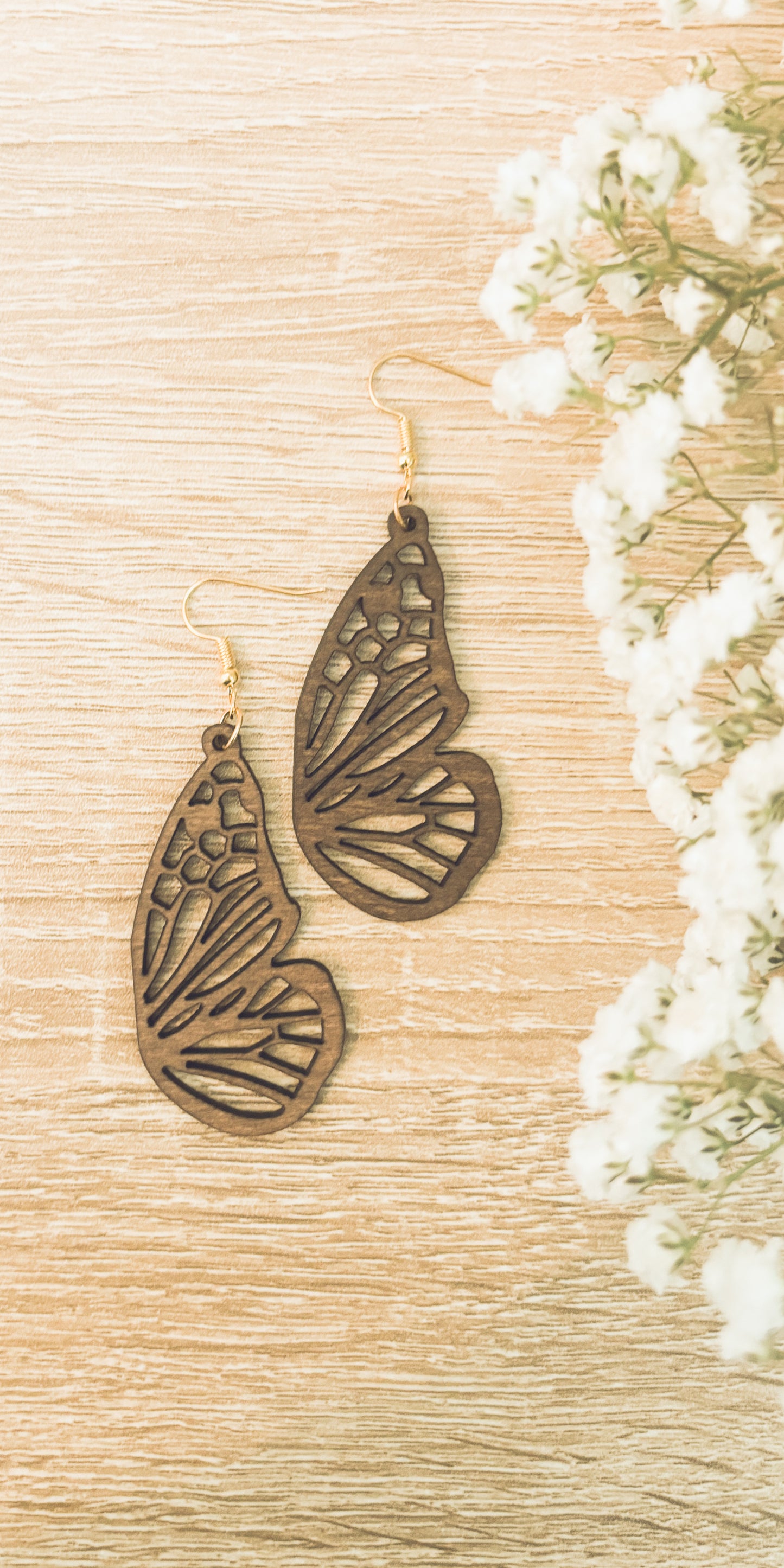 A pair of intricate laser-cut wooden butterfly wing earrings with golden hooks displayed on a light wood surface alongside delicate white baby's breath flowers
