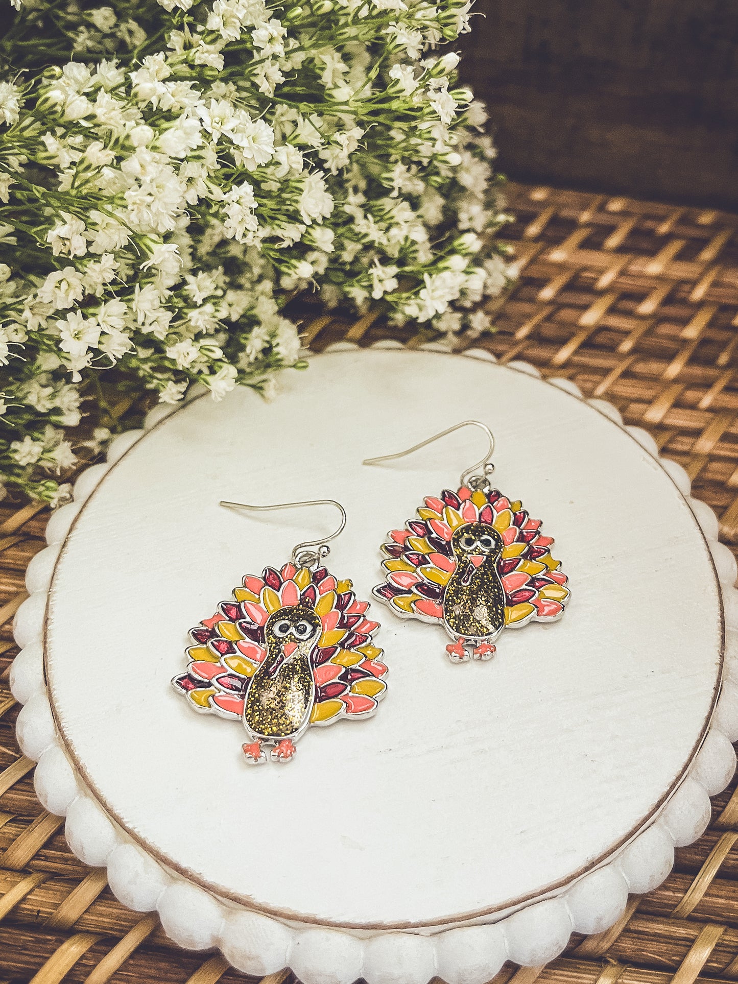 A pair of festive turkey-shaped dangle earrings with glittery brown bodies and vibrant red, pink, and yellow enamel feathers resting on a white scallop-edged display stand with blurred white flowers in the background