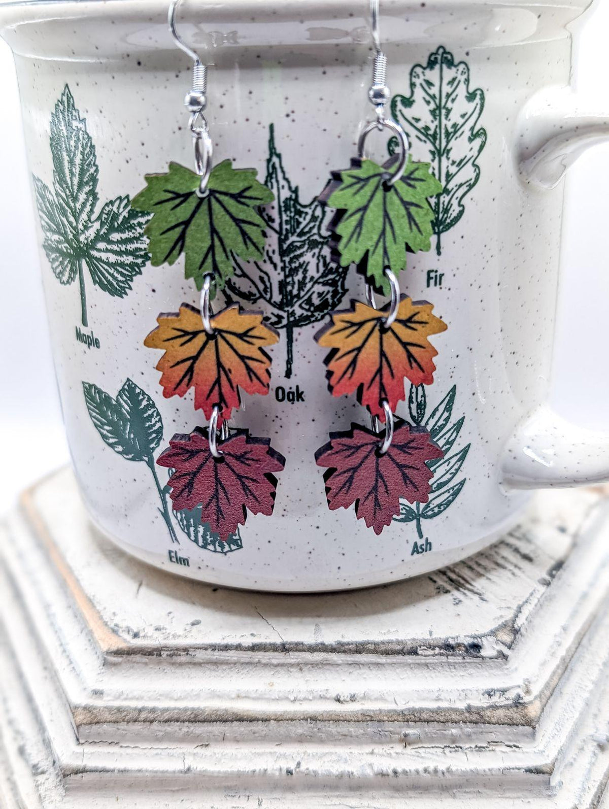 Wooden dangle earrings with silver fishhook findings, featuring tiered maple leaves in vibrant green, orange-yellow, and deep red, displayed against a speckled white ceramic mug adorned with botanical tree leaf illustrations of Maple, Oak, and Elm