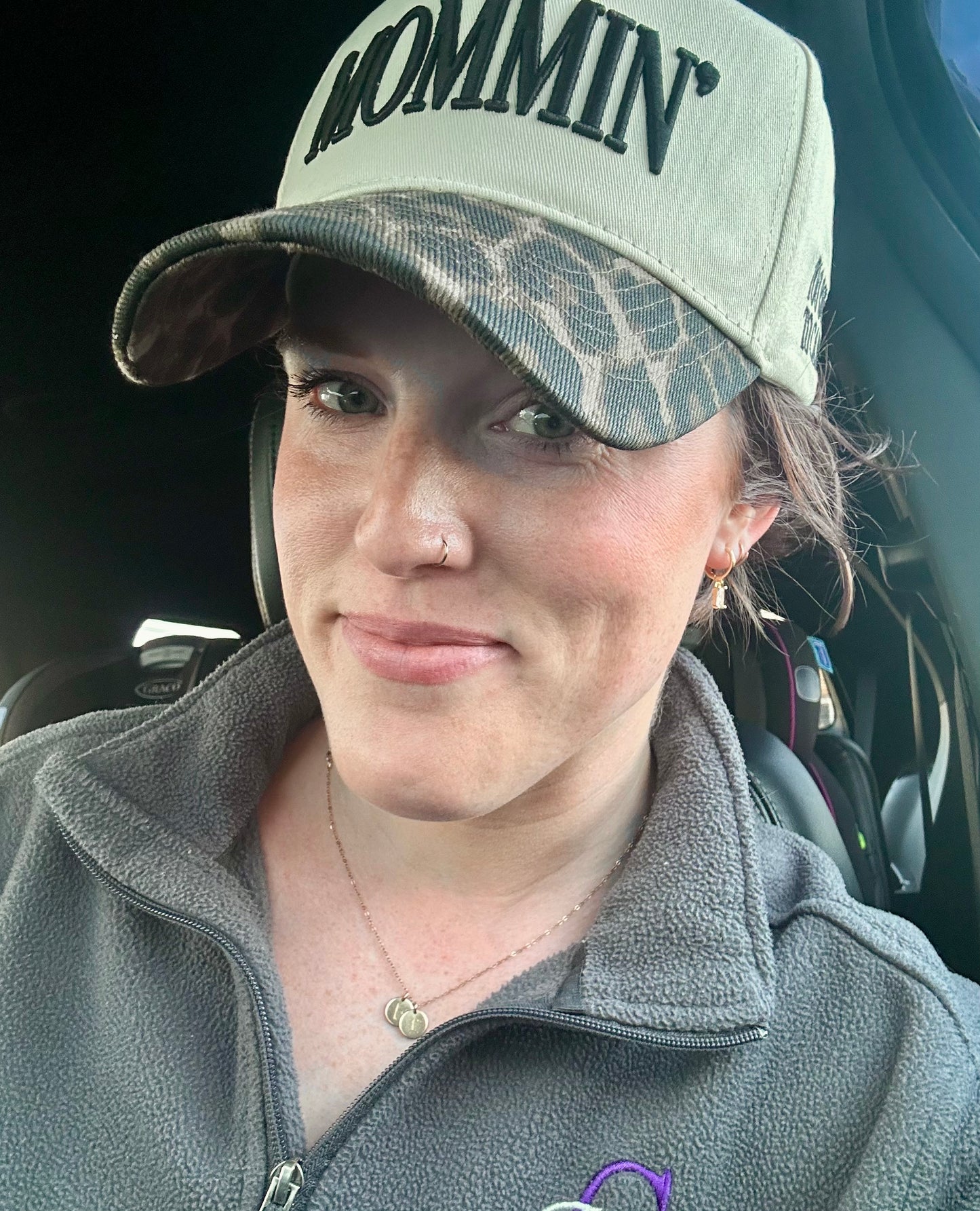 Photograph of a smiling woman with green eyes in a car wearing a light green MOMMIN' baseball cap with a camouflage brim, a small gold nose ring, and a gray fleece jacket, with a Graco baby car seat visible in the background