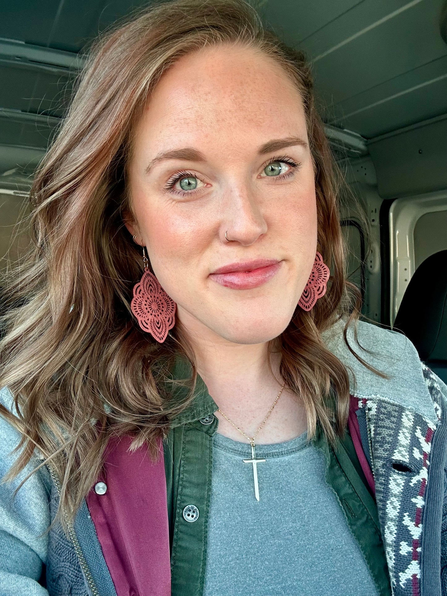 A smiling woman with striking green eyes, freckles, and a subtle nose ring, wearing ornate pink patterned earrings and a silver cross necklace, inside a vehicle