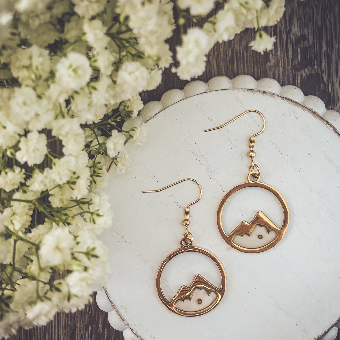 Photograph of two elegant gold-toned circular dangle earrings featuring a stylized mountain silhouette with white enamel peaks, artfully arranged on a distressed white scalloped tray alongside a cluster of delicate blurred white baby's breath flowers