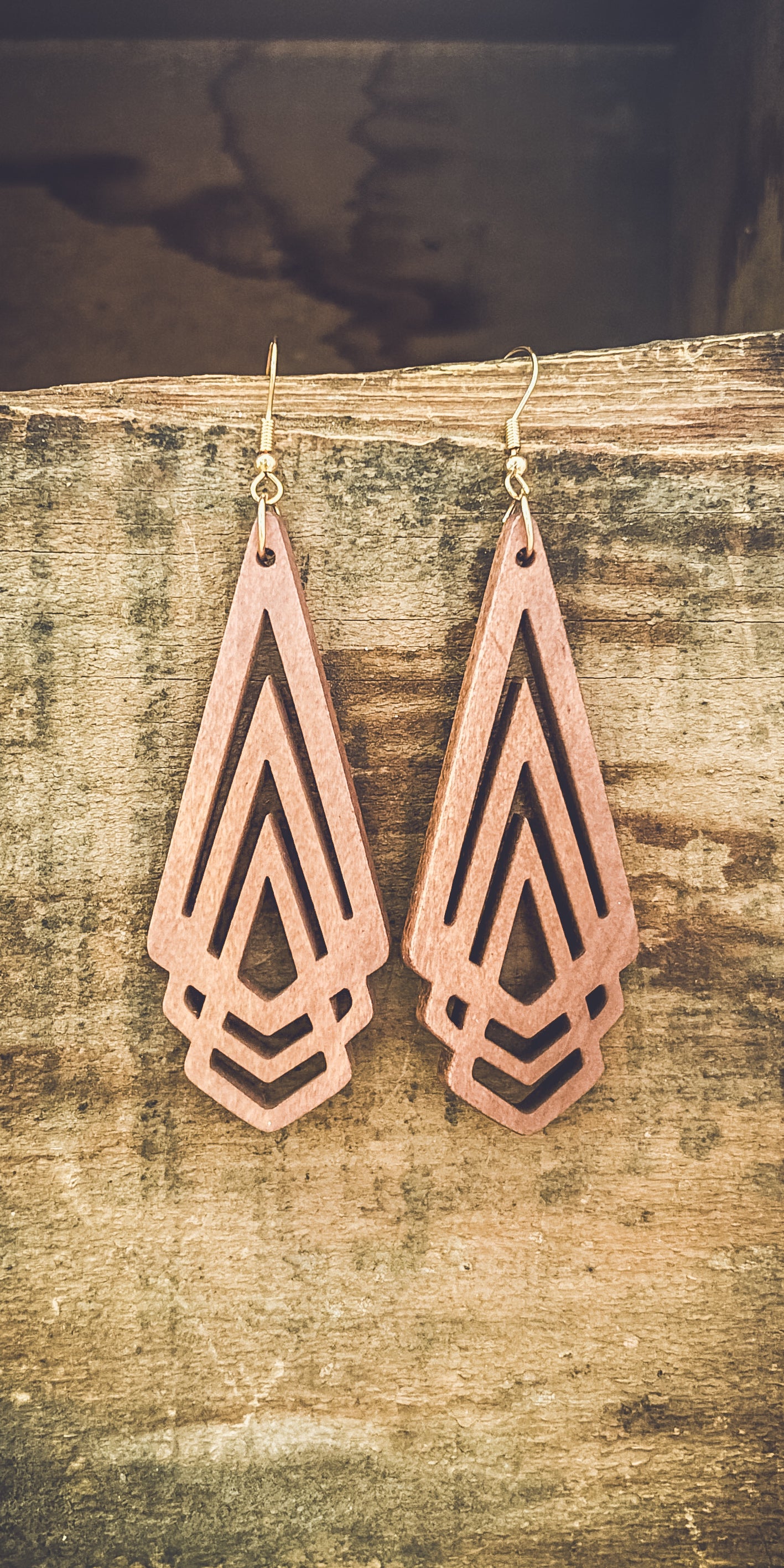 A pair of light reddish-brown geometric wooden earrings featuring layered triangle and chevron cutouts with gold hooks hanging against a rustic textured wood surface