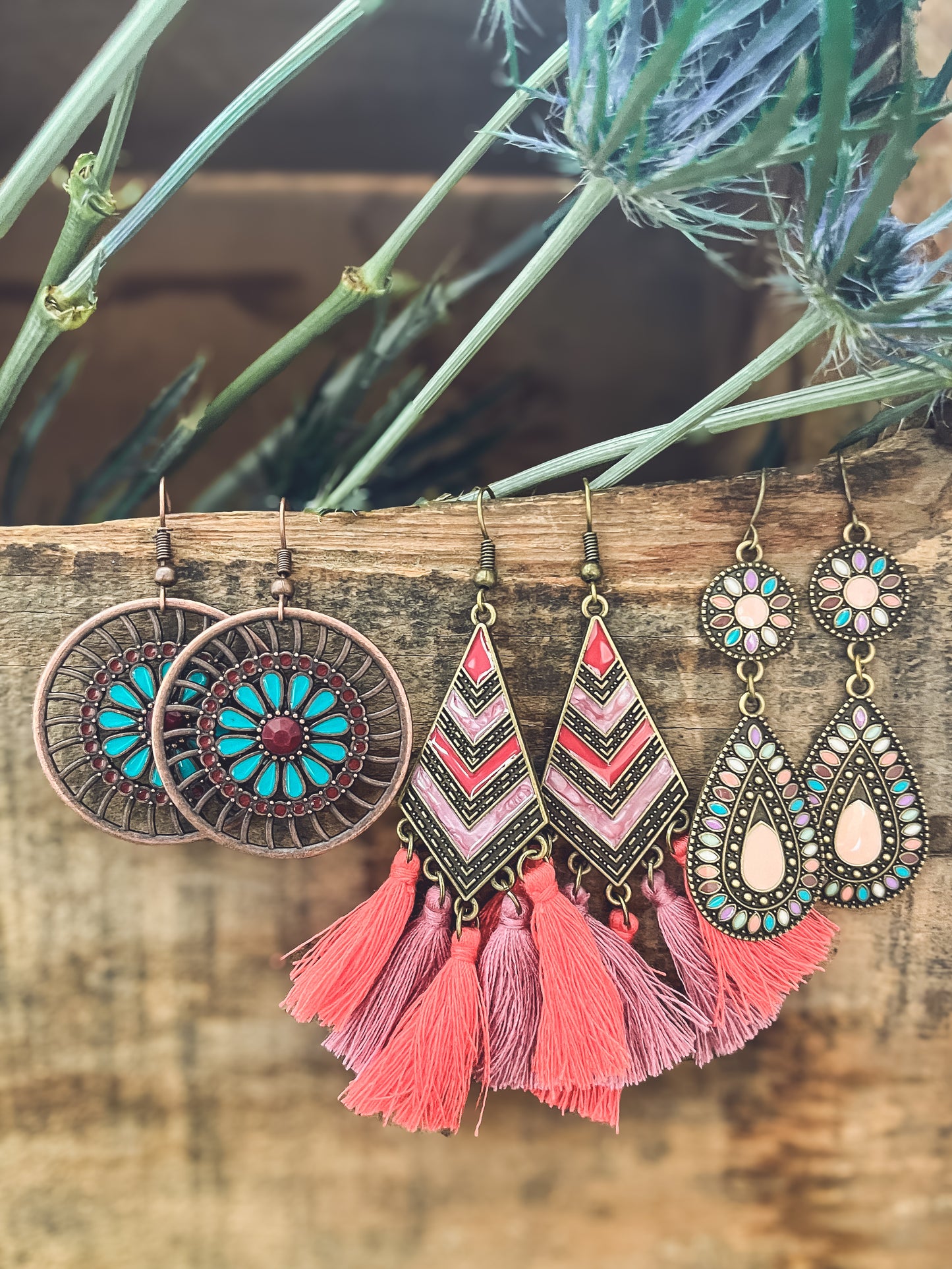 Three pairs of vibrant bohemian earrings hanging on a rustic wooden plank against a backdrop of green spiky thistles, including copper mandalas with turquoise petals, geometric pink and coral striped triangles with long tassels, and gold teardrops adorned with colorful pastel enamel