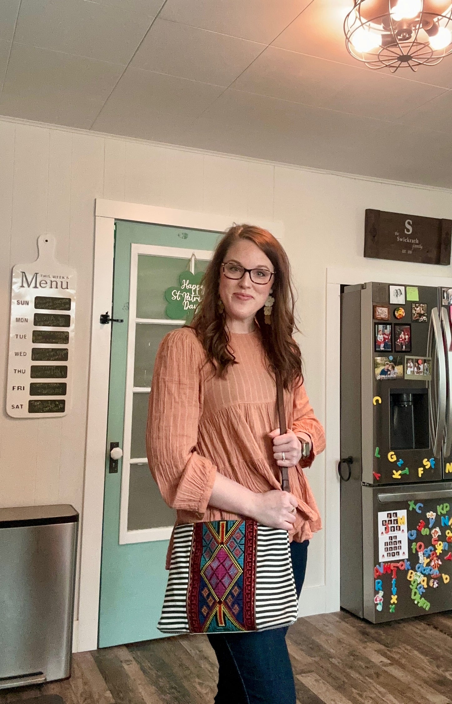 Photograph of a smiling woman with red hair and glasses wearing a peach top and carrying a striped bag with colorful geometric embroidery in a kitchen featuring a turquoise door with a 'Happy St Patrick's Day' sign and a refrigerator covered in magnets